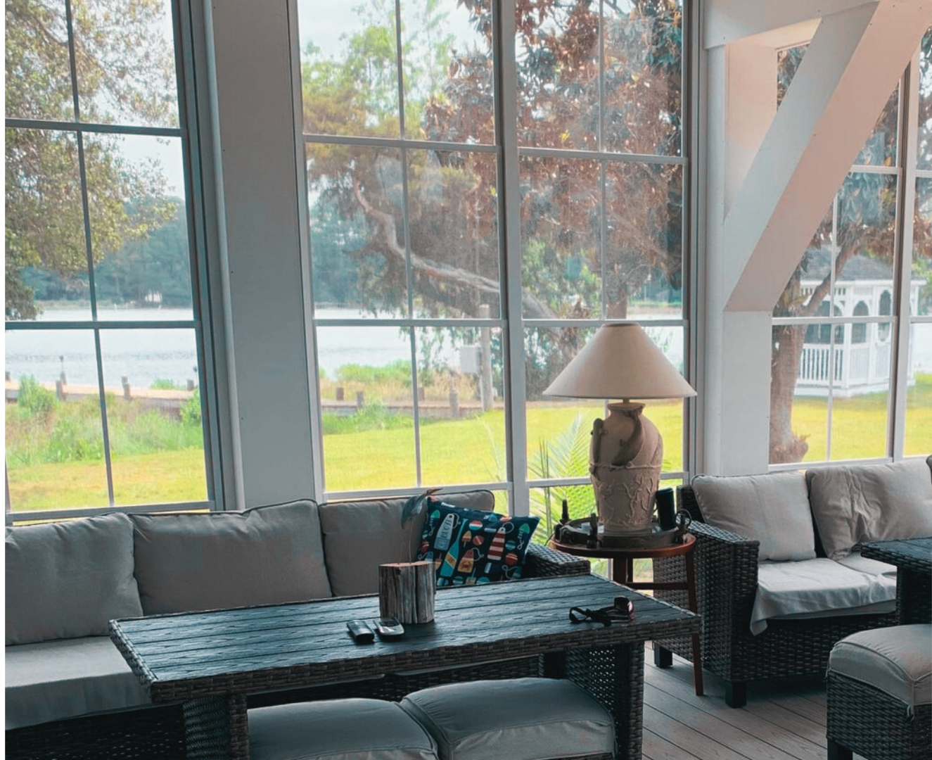 Sunroom with wicker furniture, large windows overlooking water and trees.