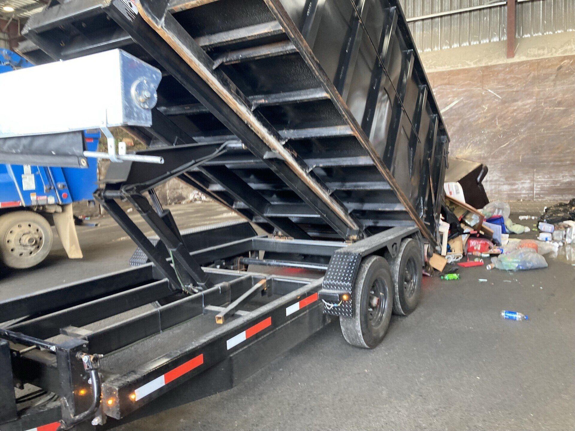 Black dump trailer with raised bed at a roadside cleanup site, next to piles of trash