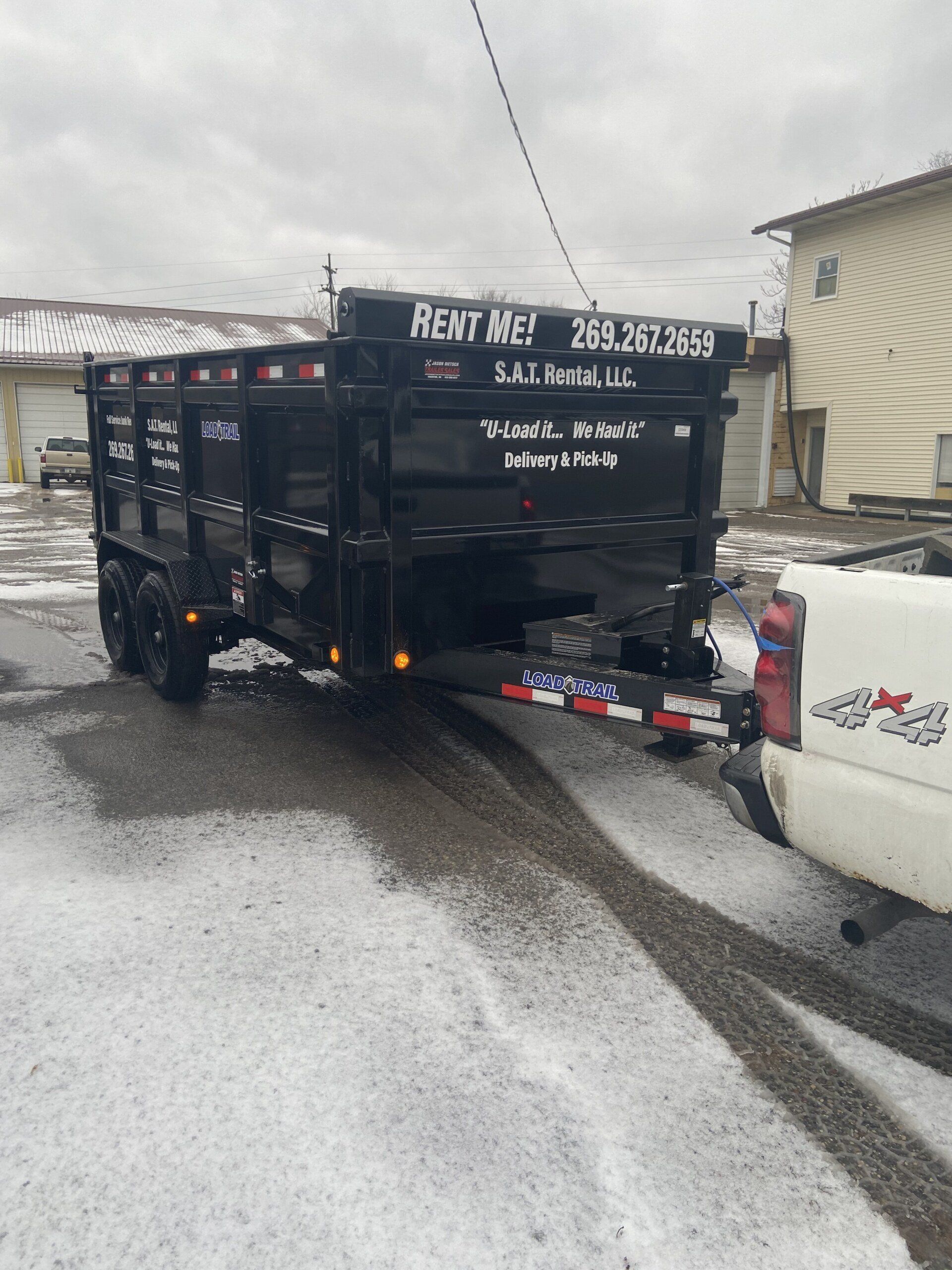 Black utility trailer behind white pickup on snowy road with debris scattered nearby