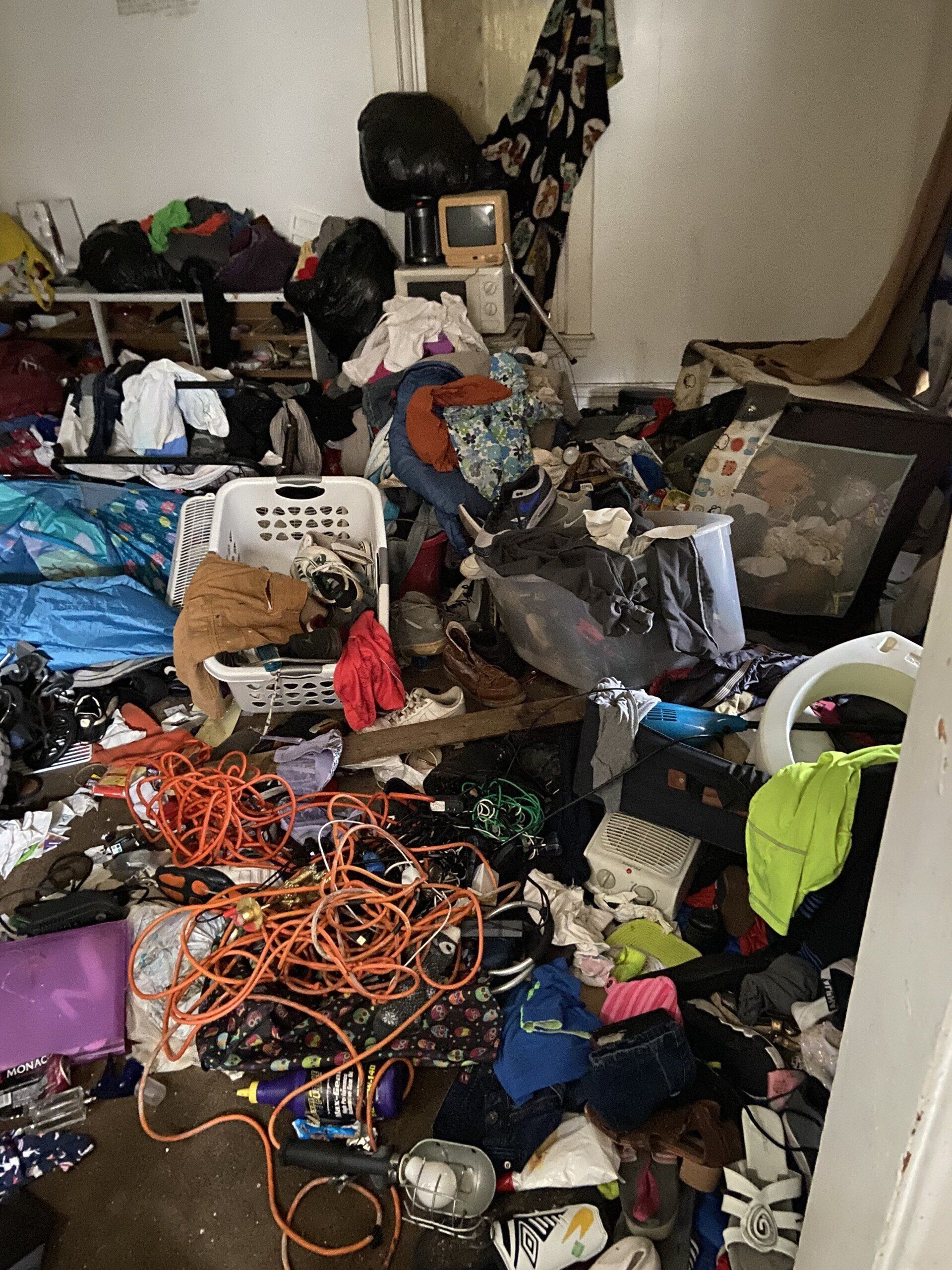 Cluttered room filled with piled clothes, cables, bags, and household items in disarray.