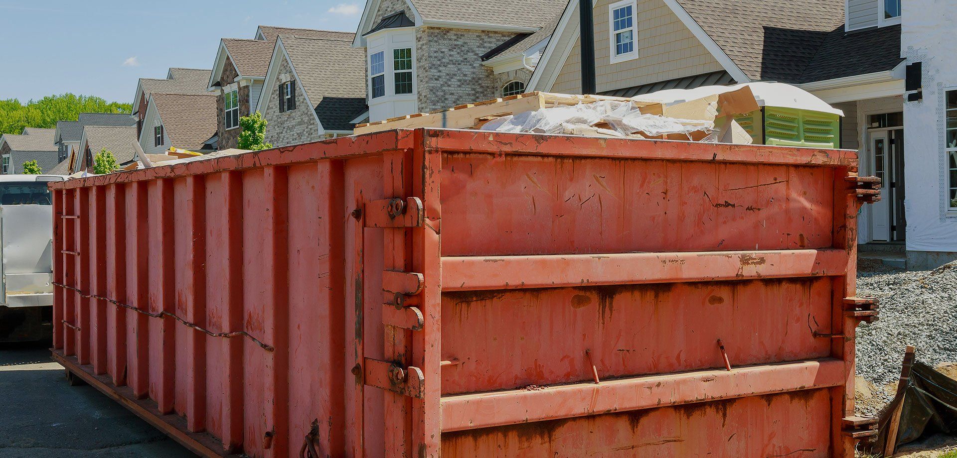 Large red construction dumpster on a gravel lot in front of houses