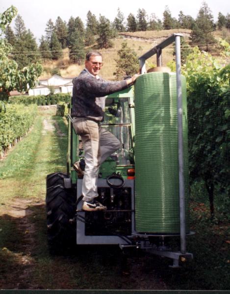 Man standing on a tractor, spraying rows of grape vines in a vineyard. Green foliage and rolling hills in the background.