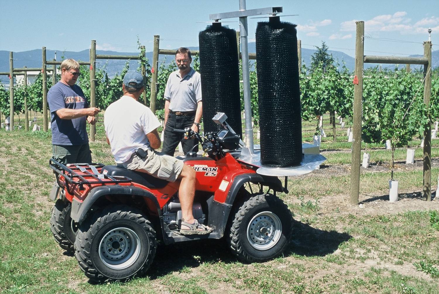 Three men with an ATV, vineyard setting. A machine with two black brushes is mounted on the vehicle.