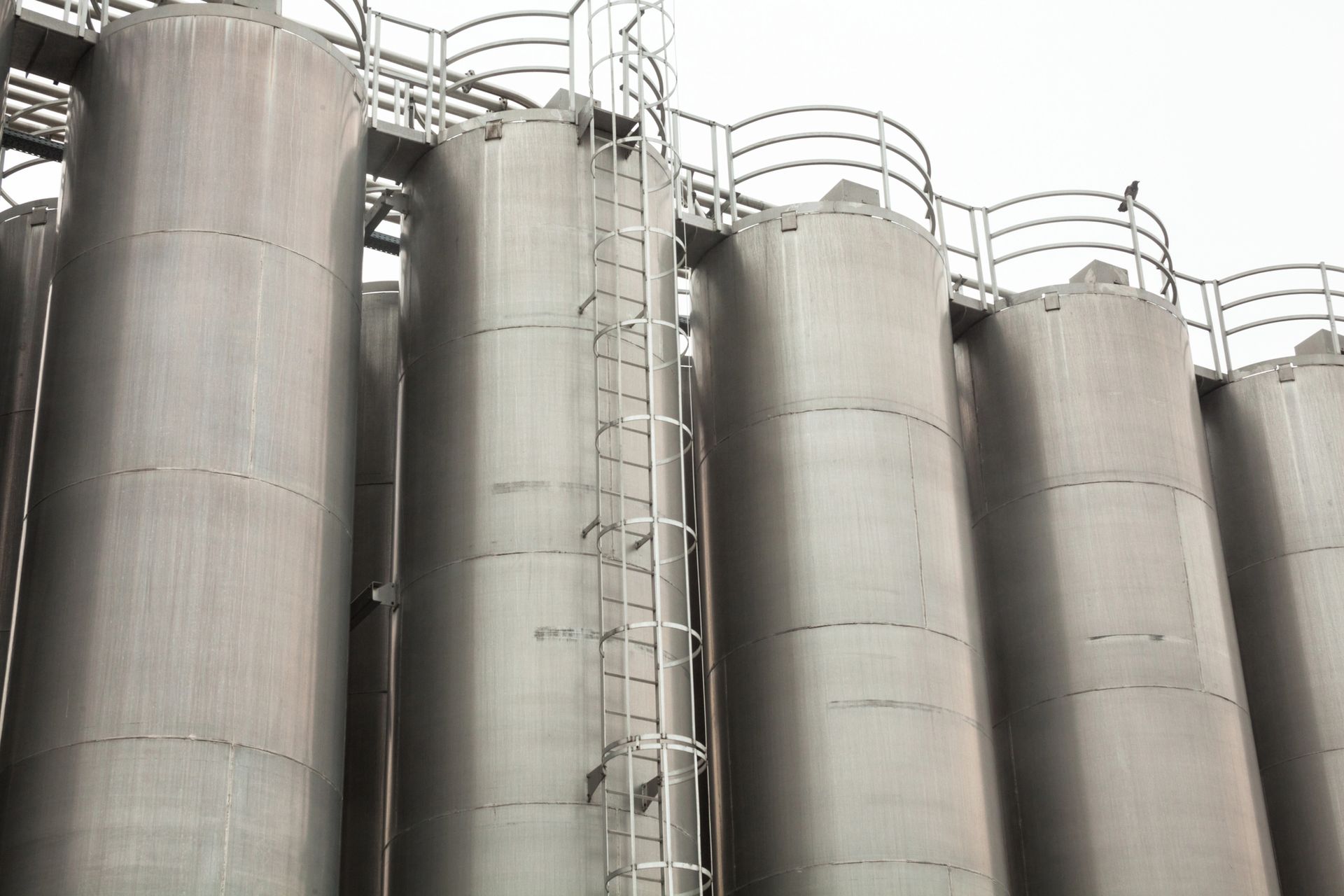 Serbatoi di stoccaggio industriali in metallo disposti in fila, con scale e passerelle, contro un cielo nuvoloso.