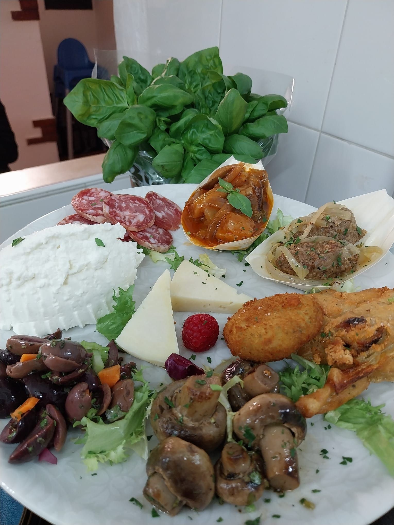 Un assortimento di piatti mediterranei con riso, olive, funghi, crocchette e verdure a foglia verde, disposti su un tavolo bianco.