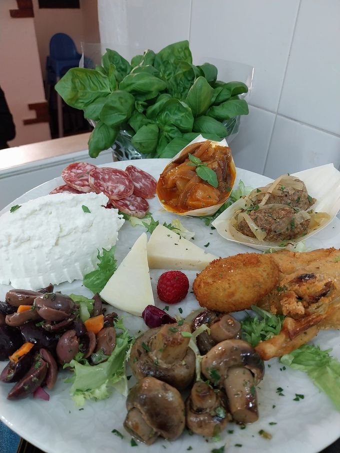Un assortimento di piatti mediterranei con riso, olive, funghi, crocchette e verdure a foglia verde, disposti su un tavolo bianco.
