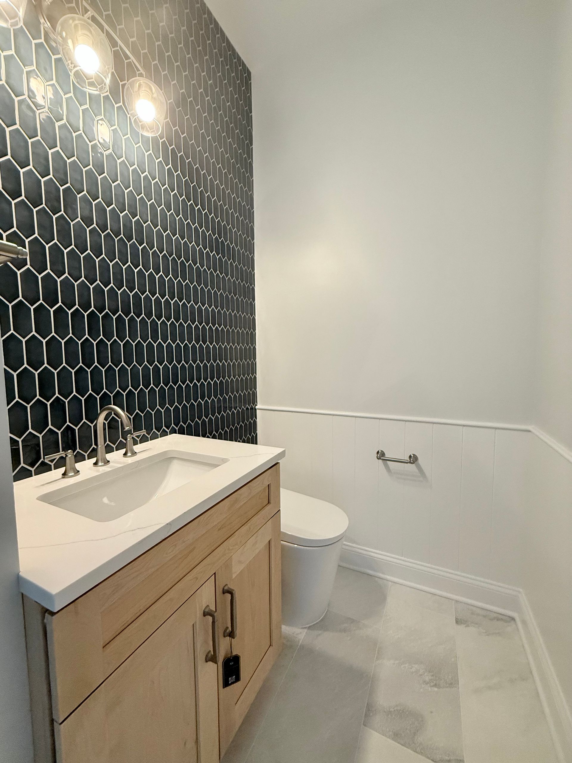 A full view of a powder room remodel showing the bold hexagonal tile accent wall, a natural wood vanity, and a new smart toilet.