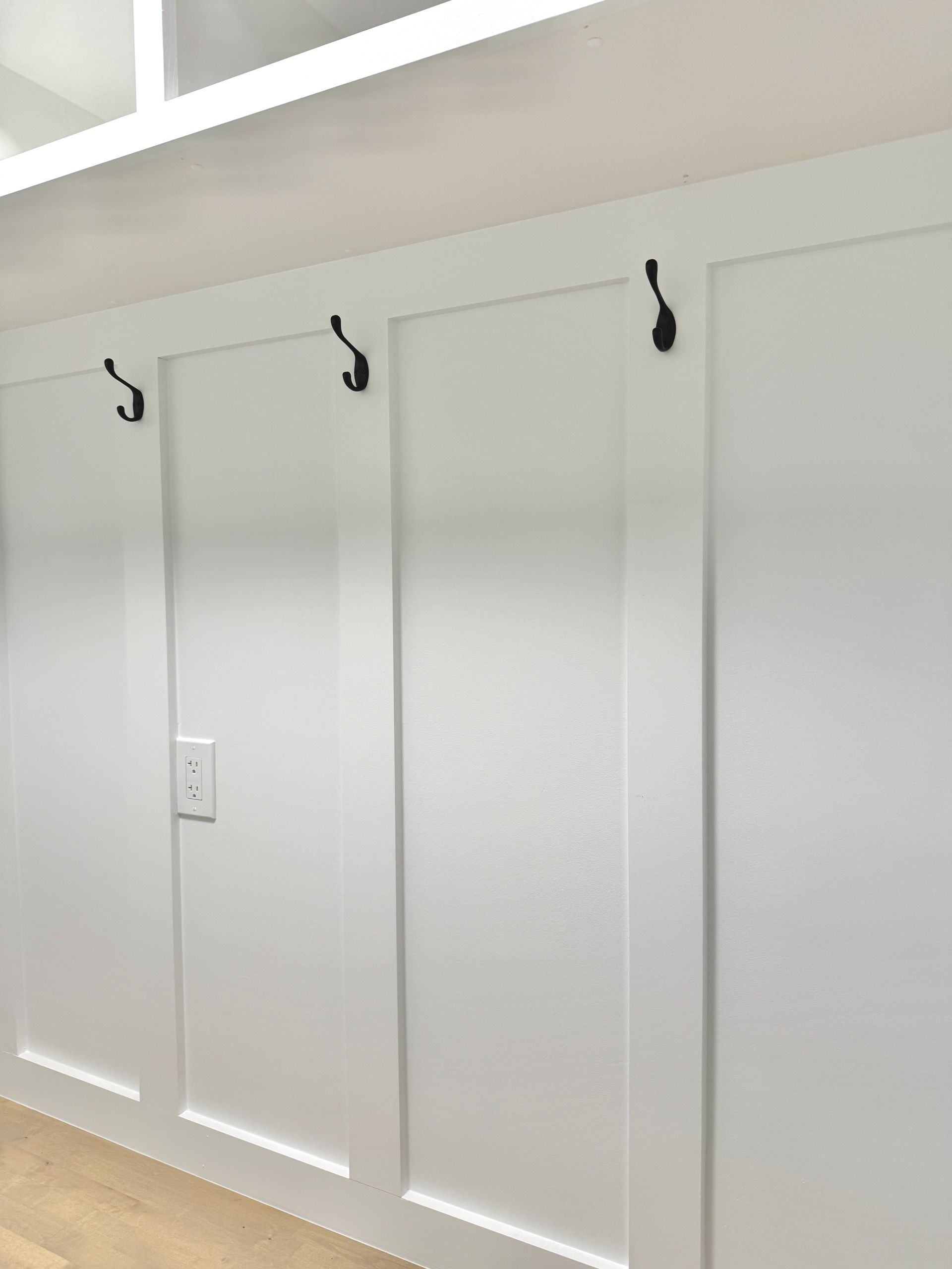 Detail of matte black coat hooks on custom white board-and-batten paneling with electric outlet.