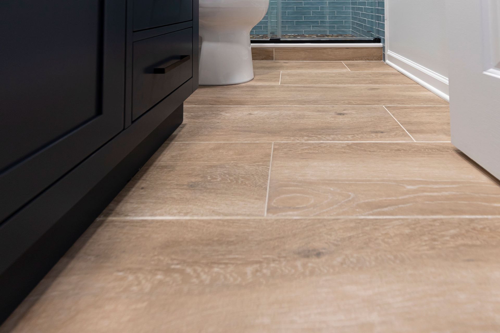 Wood-look tile floor and the base of the modern black vanity near the shower entrance in the renovated bathroom.