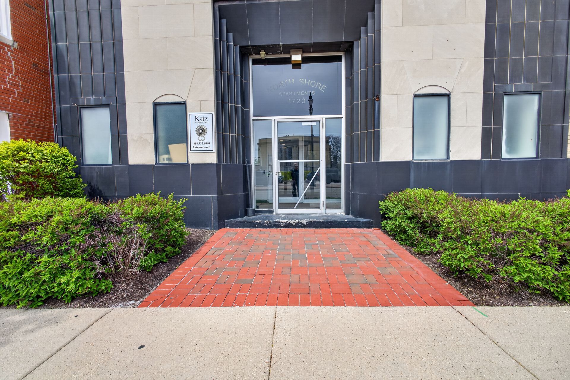 Building entrance with glass doors, red brick walkway, and shrubs on both sides