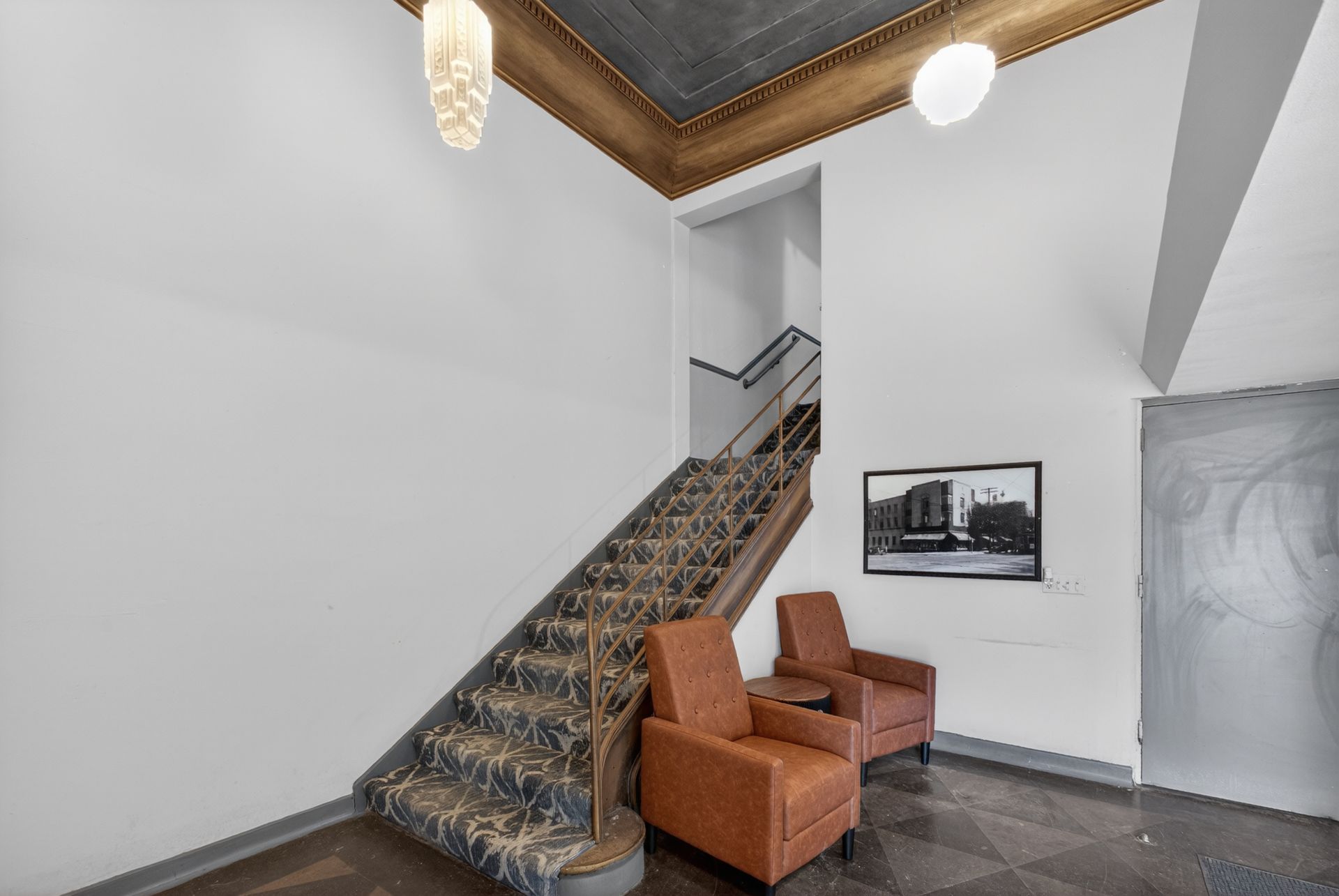 Interior staircase with patterned carpet, orange chairs, framed wall art, and a high ceiling with a chandelier.