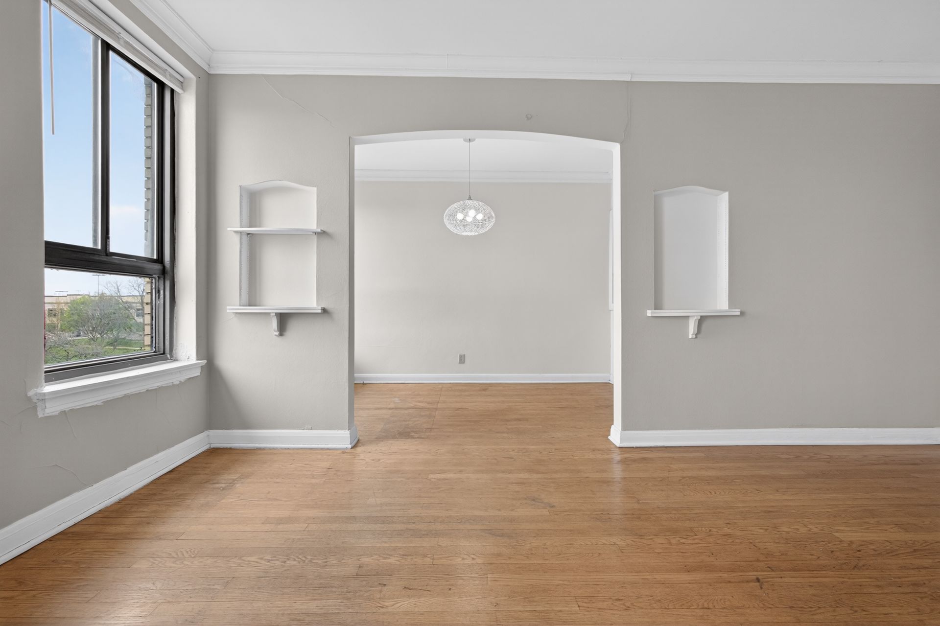 Empty room with hardwood floors, gray walls, a large window, and a doorway into another room.
