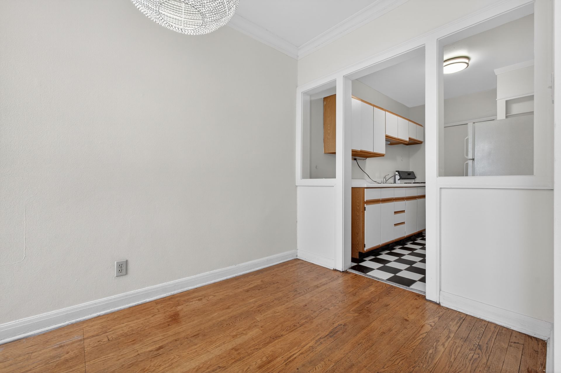 Empty dining area with hardwood floor, leading to a white kitchen with black-and-white checkered tile.