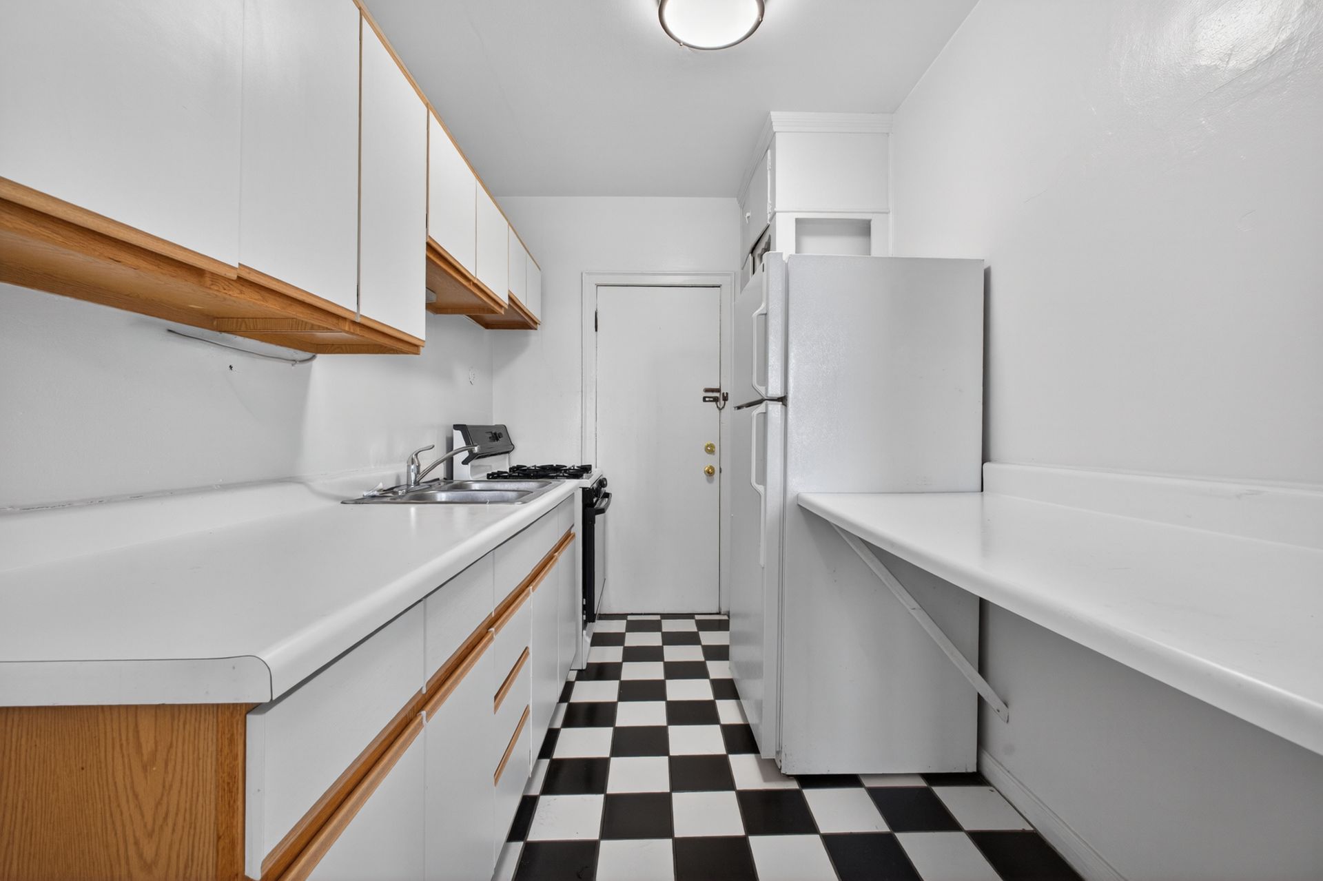 Narrow white kitchen with black-and-white checkered floor, long counters, cabinets, sink, and refrigerator.