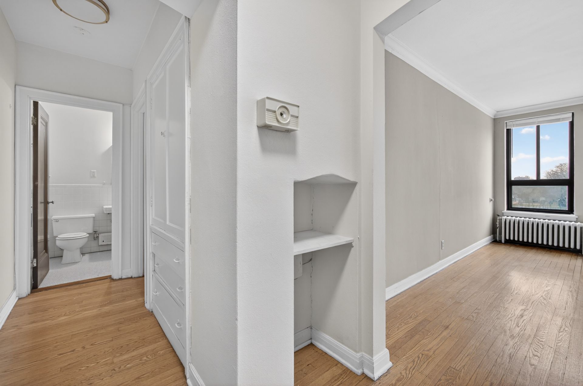 Bright hallway and empty room with hardwood floors, gray walls, white trim, and a window with radiator underneath