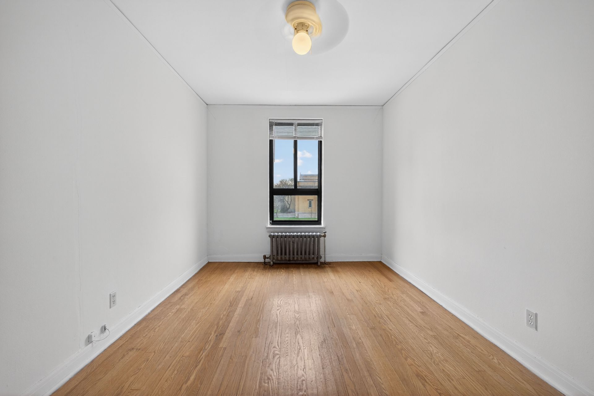 Empty white room with hardwood floors and a single window with radiator beneath it
