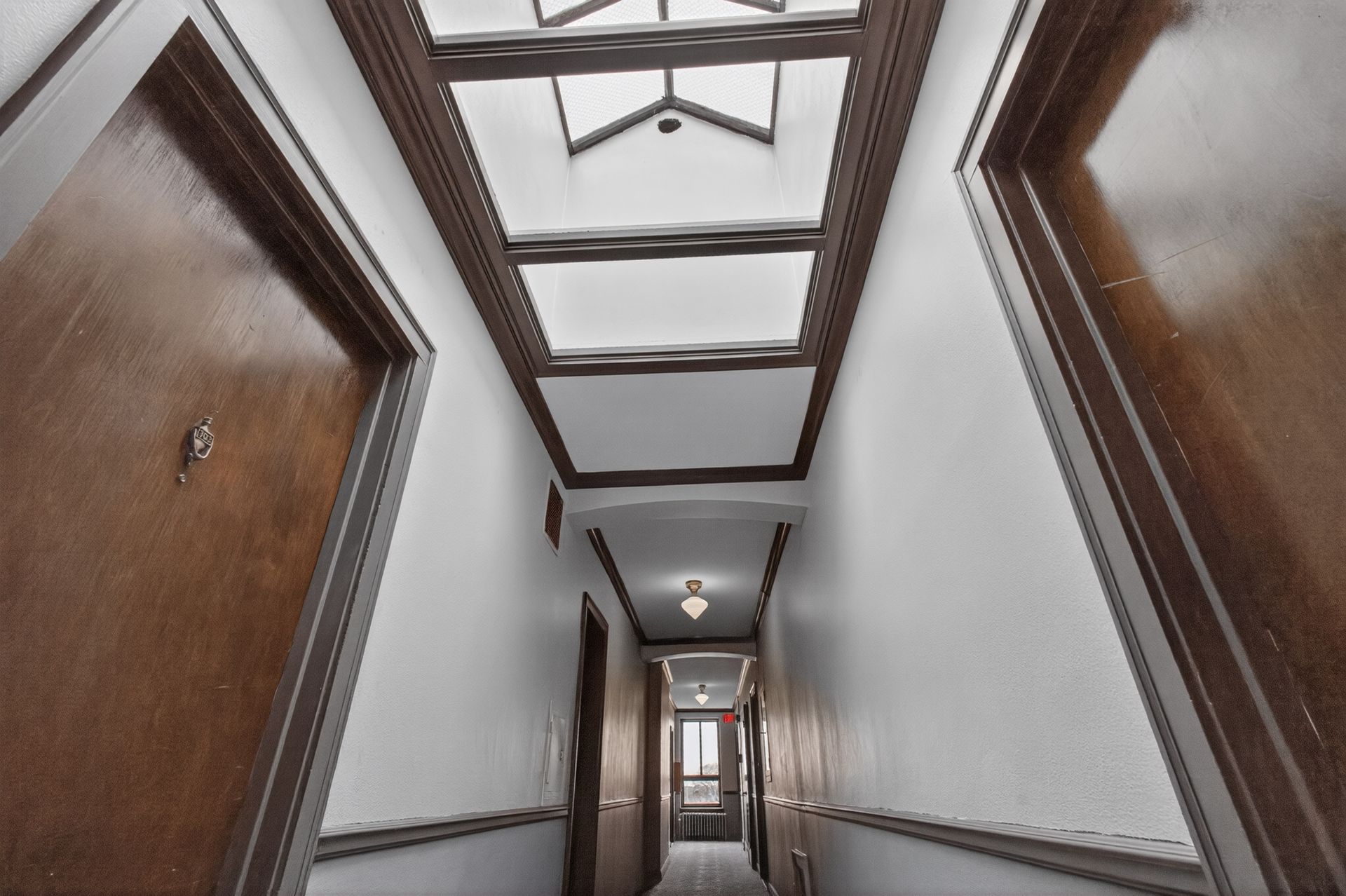 Narrow hallway with wooden doors, white walls, and exposed ceiling beams leading to a lit doorway