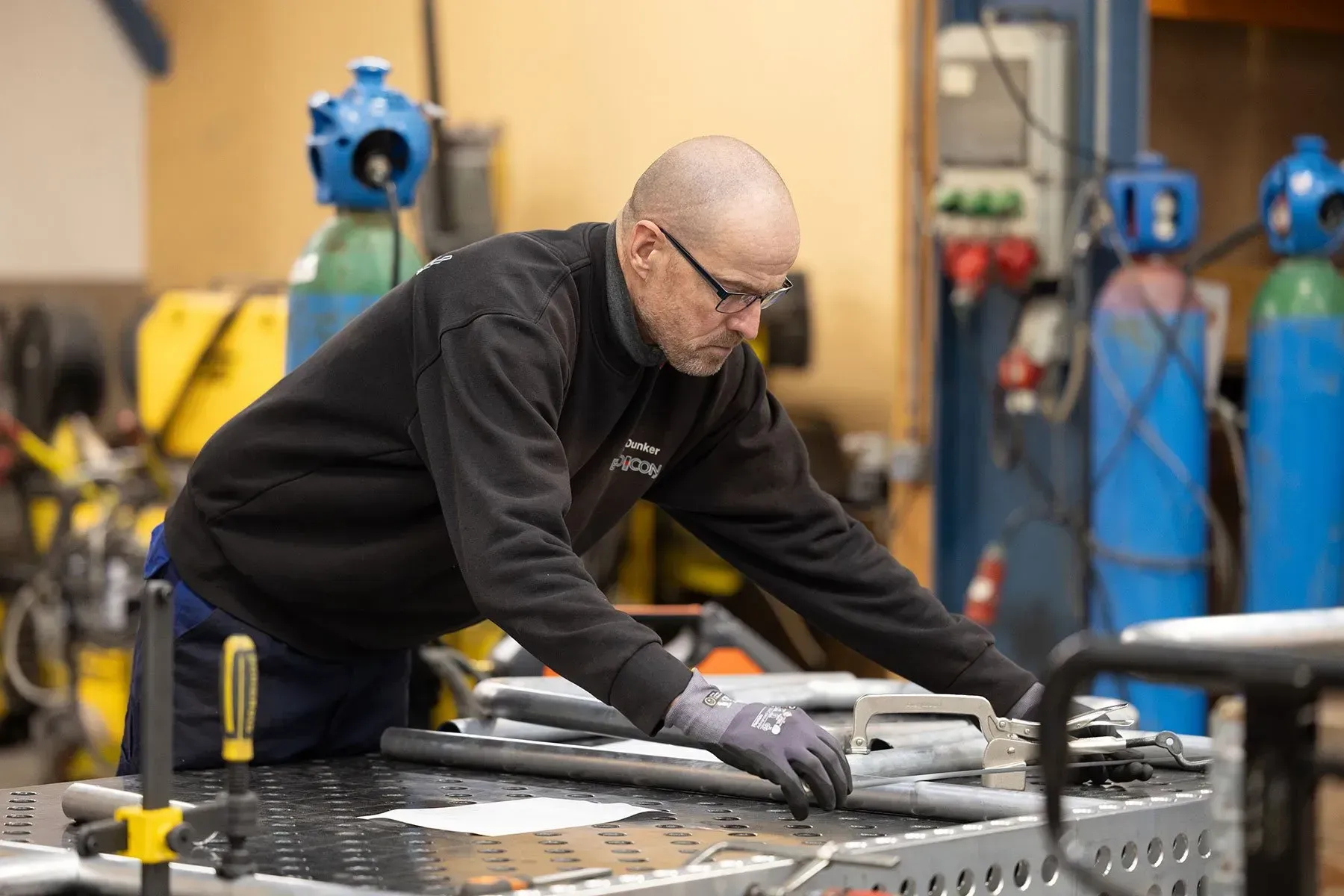 Arbejder i sort jakke og handsker bøjer sig over en metalarbejdsbænk i et industriværksted med blå benzintanke bagved.