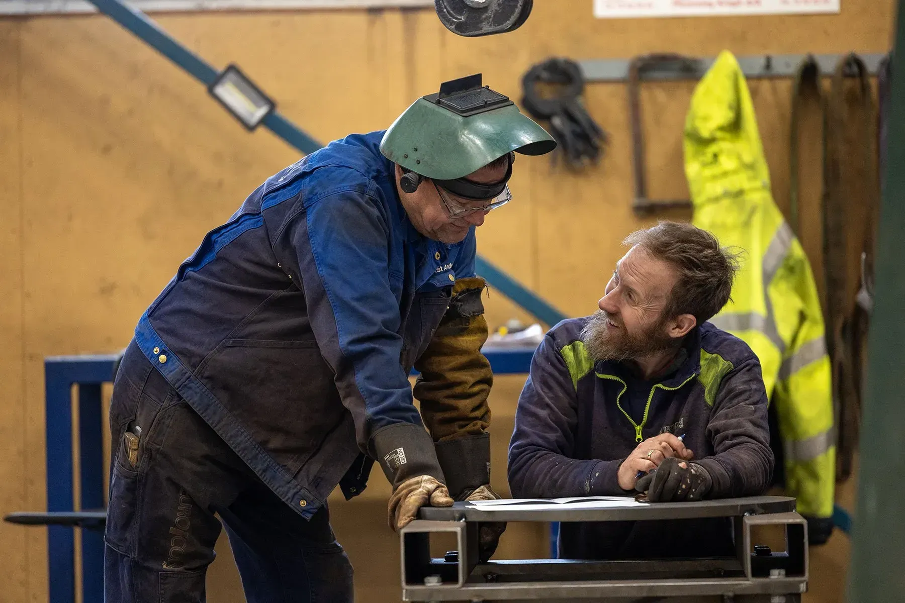 To arbejdere i et værksted, den ene læner sig over et bord mens han bærer et grønt ansigtsskærm og hjelm.