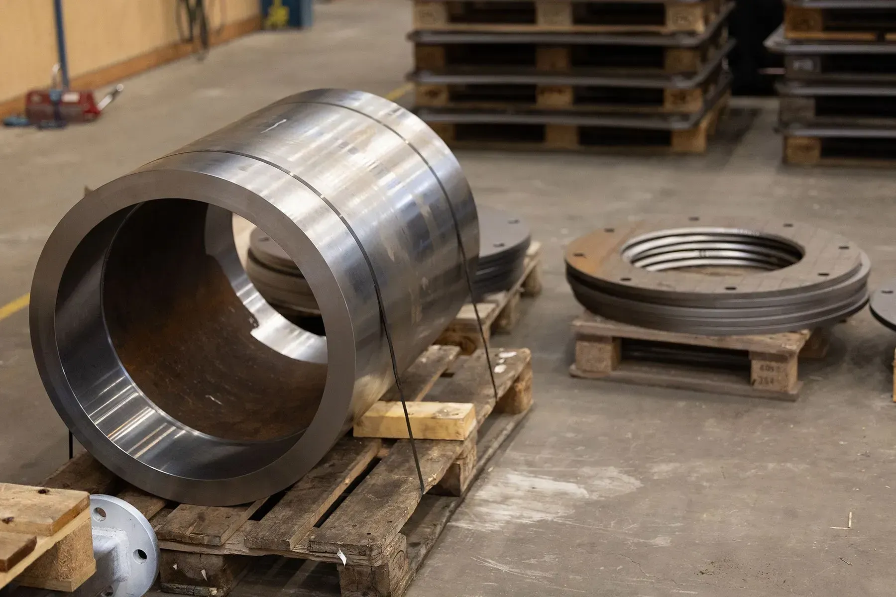 Stor stålcylinder og cirkulær metalring på træpaller i et industrielt værksted