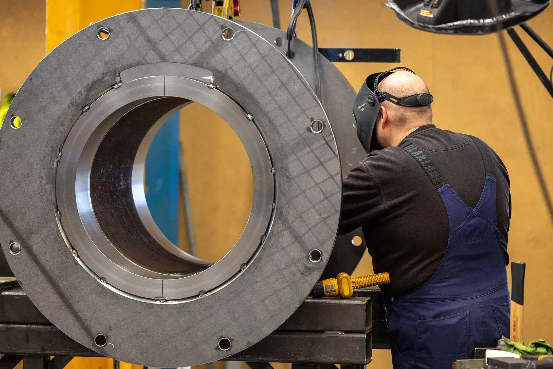 Arbejder i blå overalls inspicerer en stor metalrørflange i et industriværksted