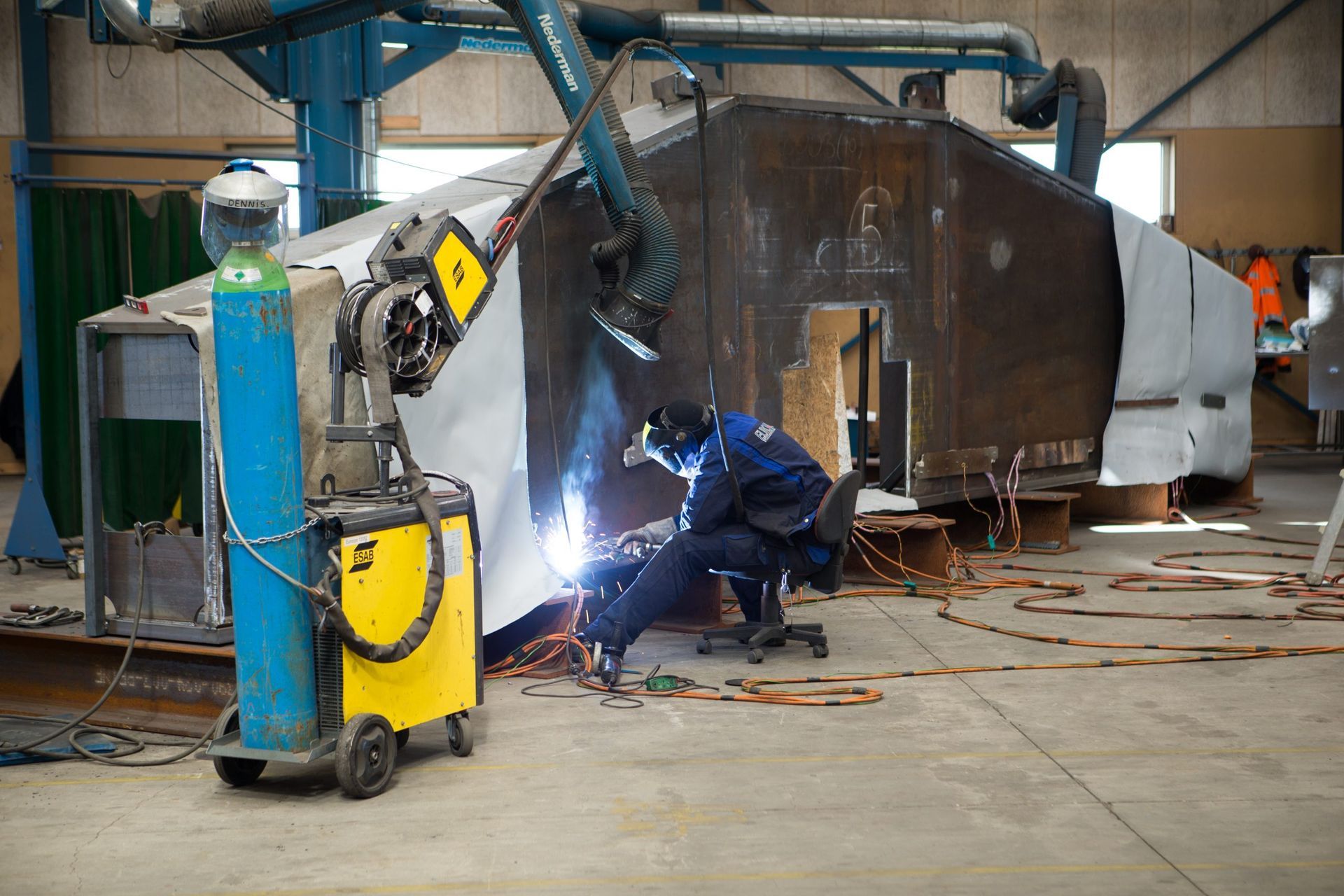 Svejser i blåt beskyttelsesudstyr gnister ved siden af ​​en stor metaltank i et industriværksted