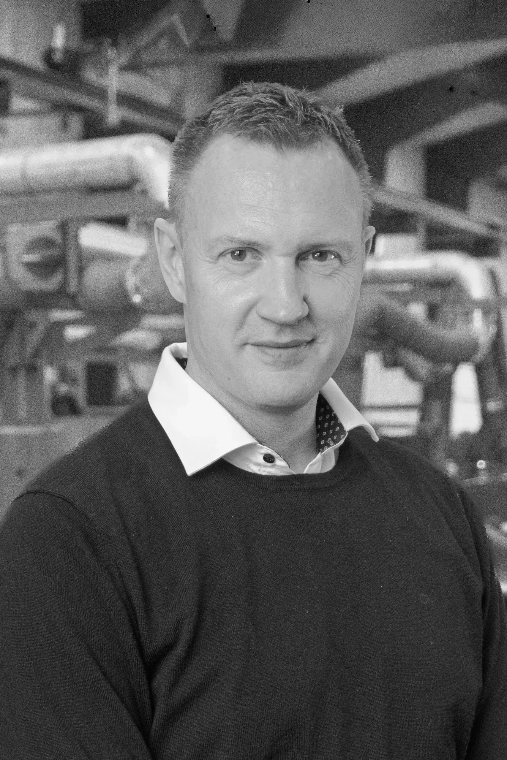 Portræt af en mand i en mørk sweater, der står i et industrielt miljø med rør bag sig