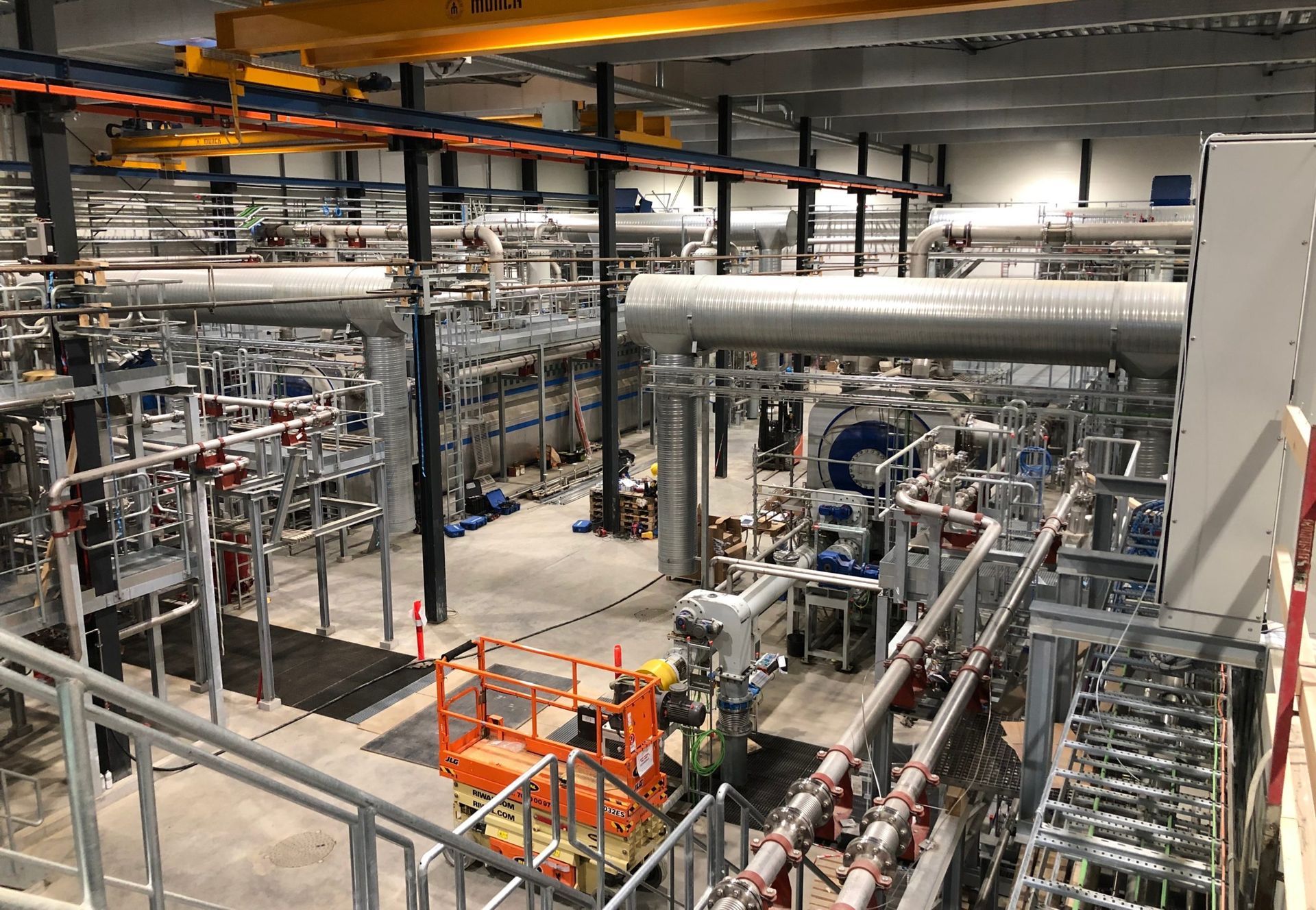 Interiør af industrianlæg med store rør, metalgangbroer og maskiner i et lyst bryggers