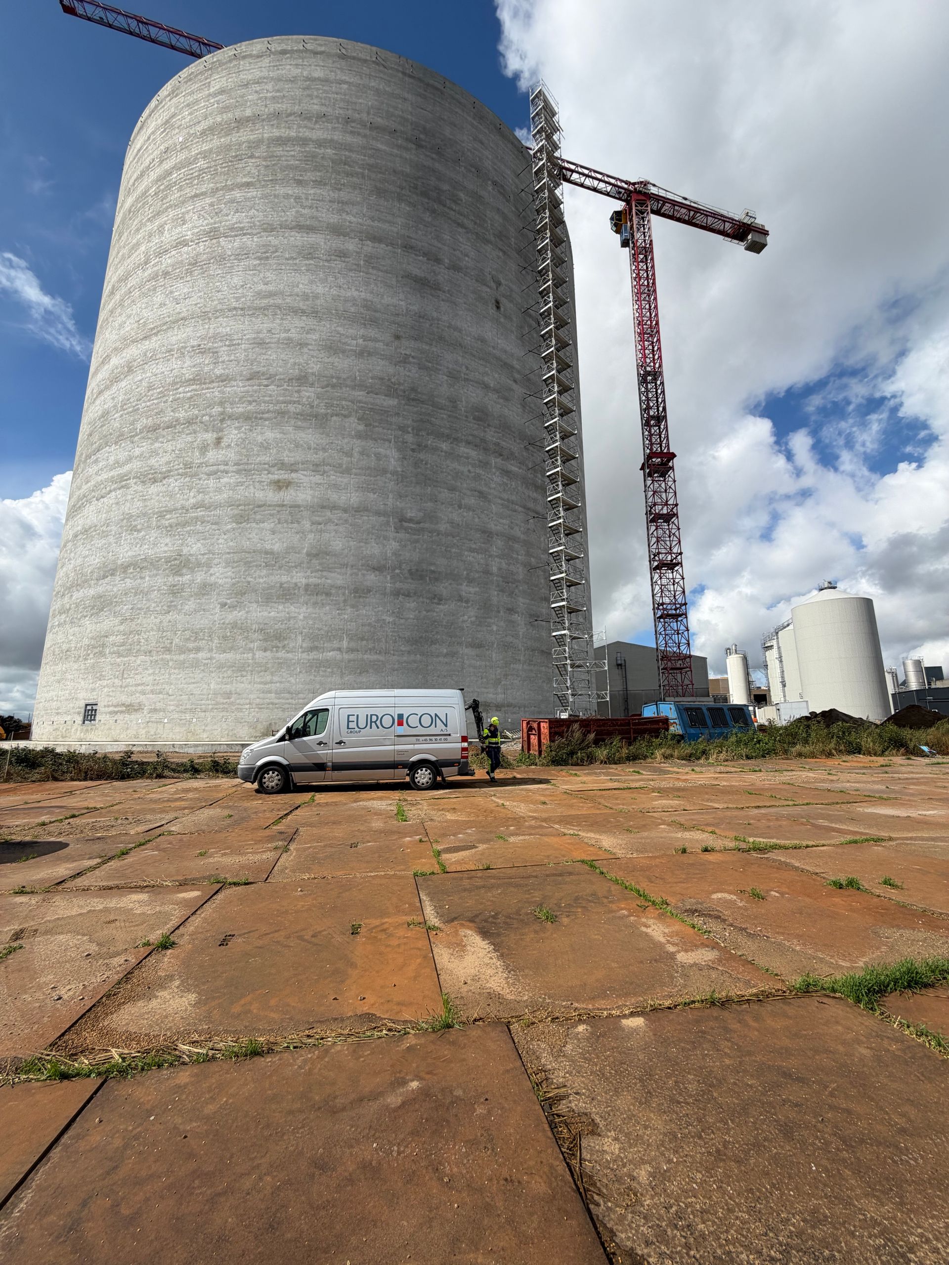 Stor betonsilo under opførelse med kran, hvid varevogn og overskyet himmel på en plads med brostensbelægning