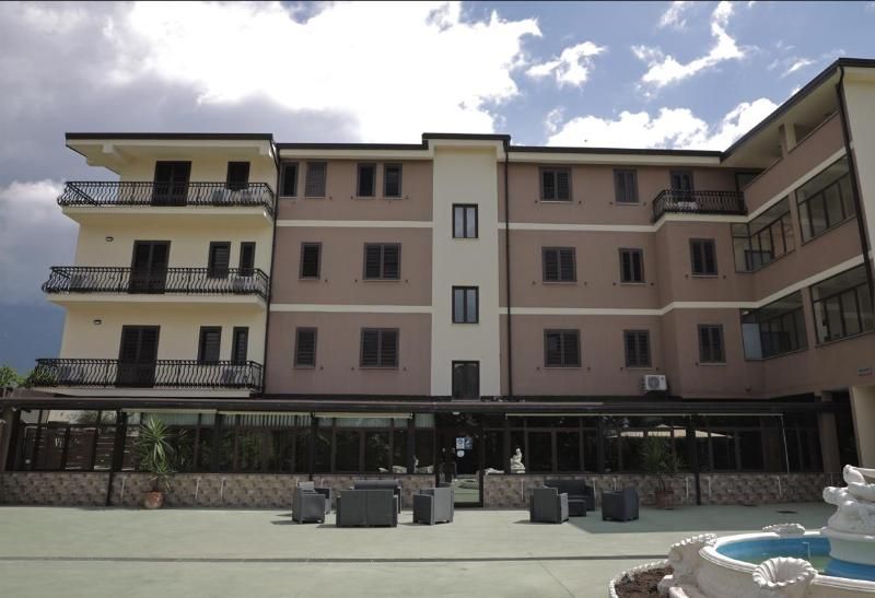 Esterno di un hotel a più piani, nei toni del beige e del bianco, con ringhiere sui balconi, posti a sedere all'aperto e una fontana in primo piano.