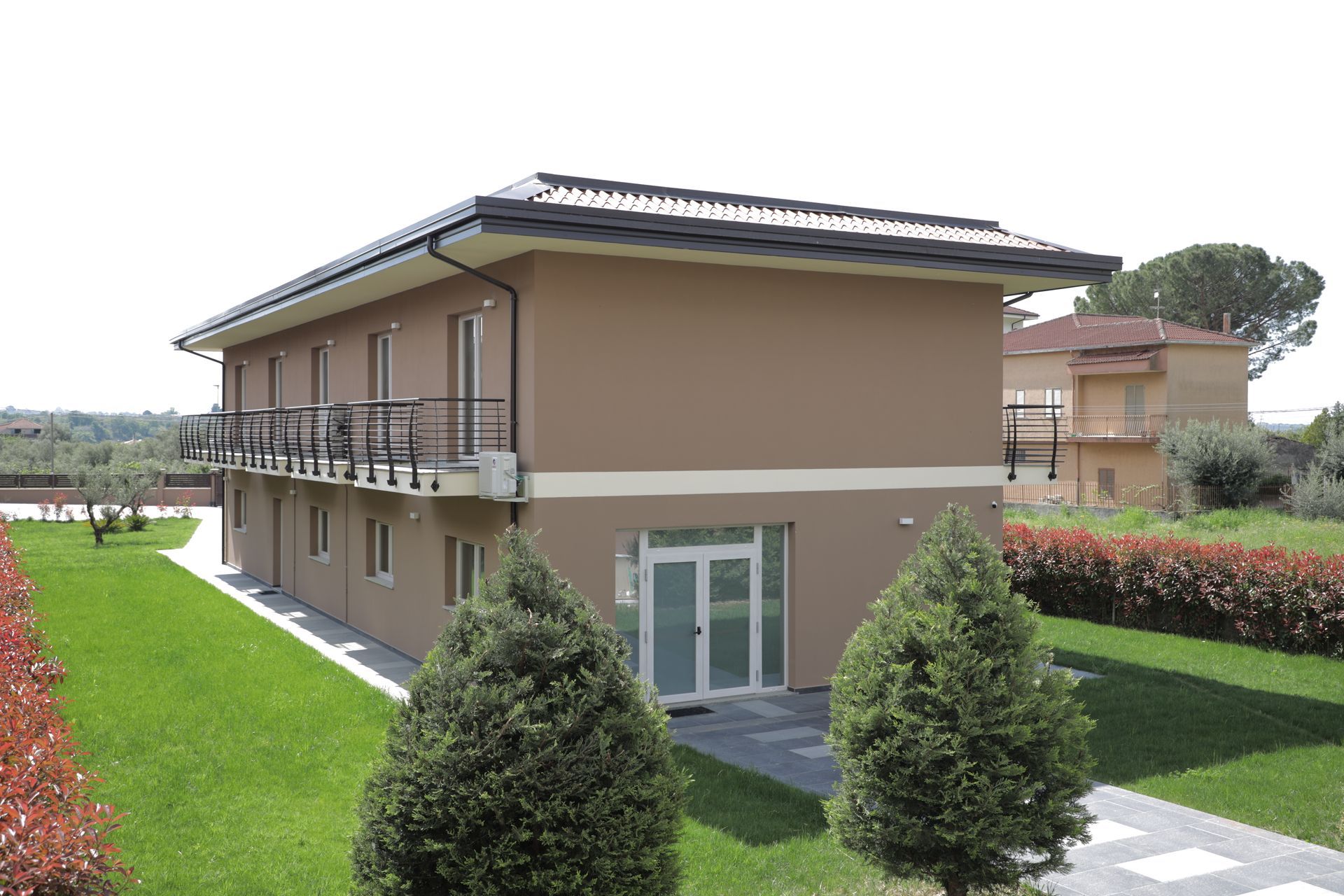 Un edificio a due piani color beige con balcone, circondato da un prato verde e arbusti ben curati sotto un cielo limpido.