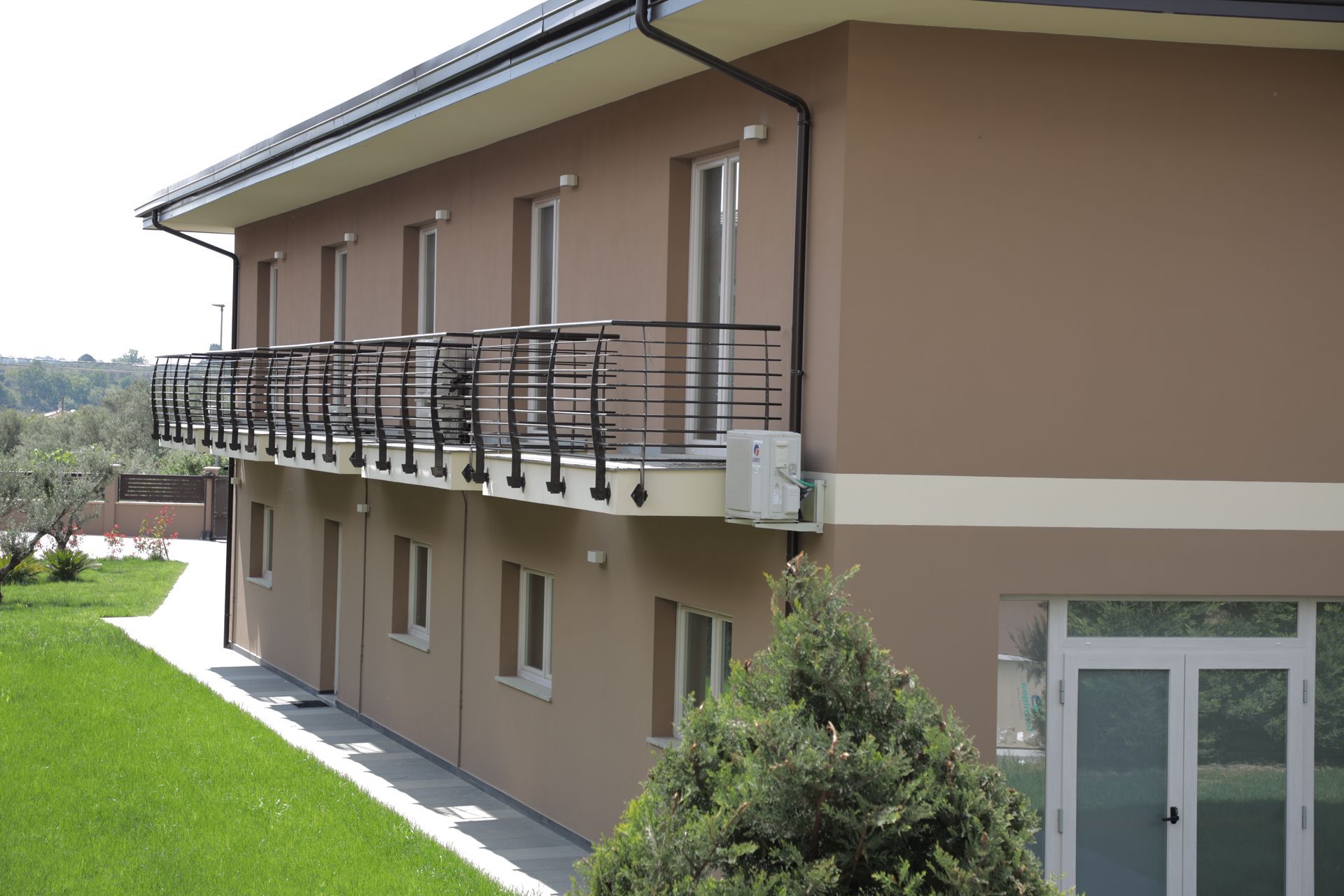 Un edificio a due piani color beige con un lungo balcone in metallo e un prato verde in primo piano.