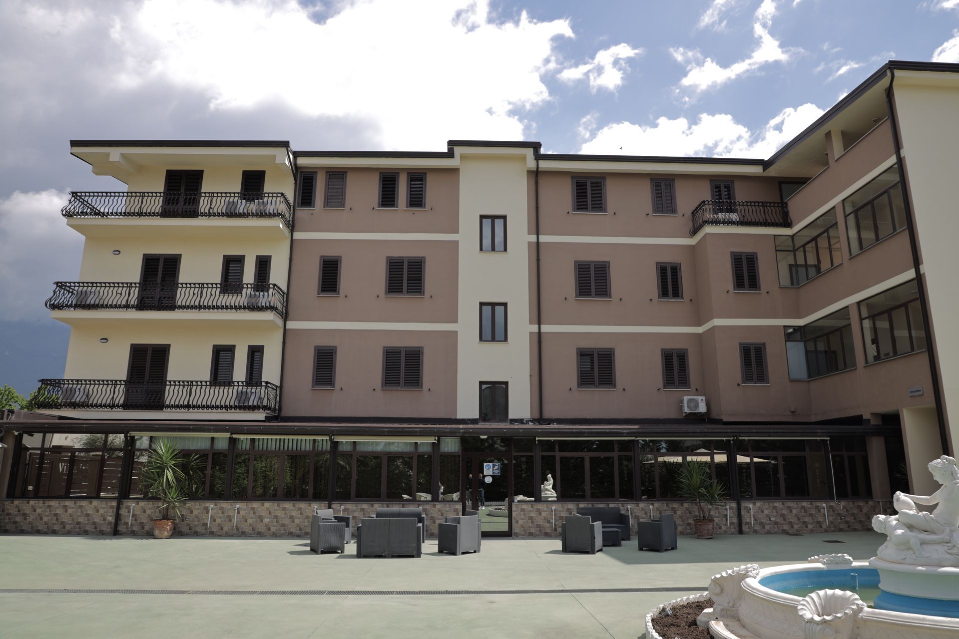 Un moderno hotel a più piani con pareti color beige, balconi, una terrazza al piano terra con mobili da esterno e una fontana in pietra.