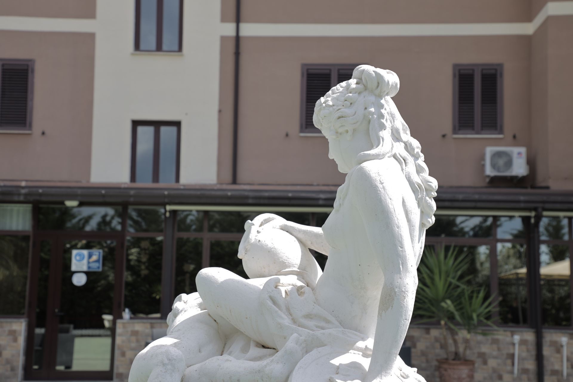 Una statua-fontana in pietra bianca raffigurante una figura che regge un vaso, situata all'aperto di fronte a un edificio a più piani di colore beige.