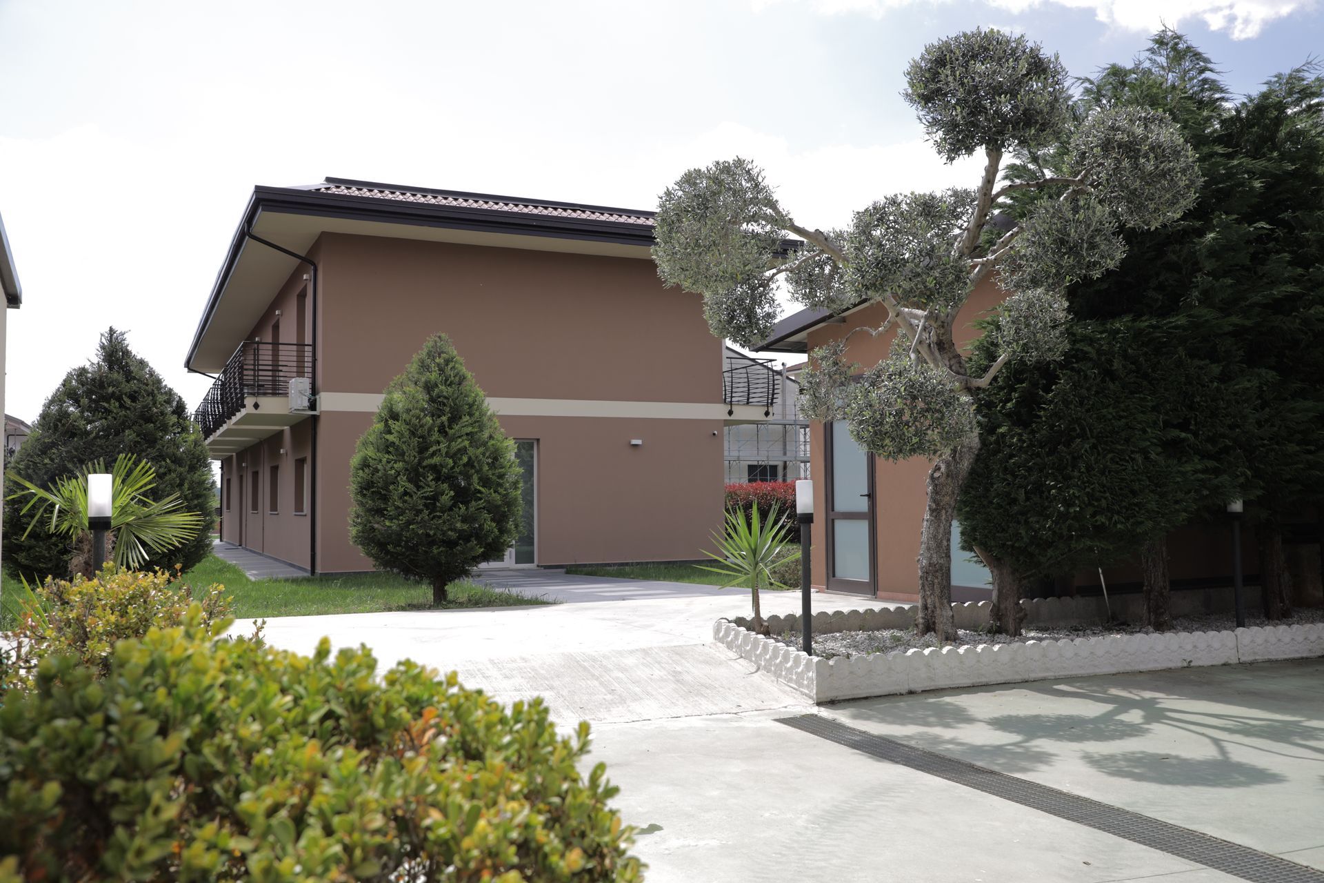 In una giornata di sole, un edificio a due piani color beige con balcone, immerso tra arbusti verdi e un vialetto lastricato.