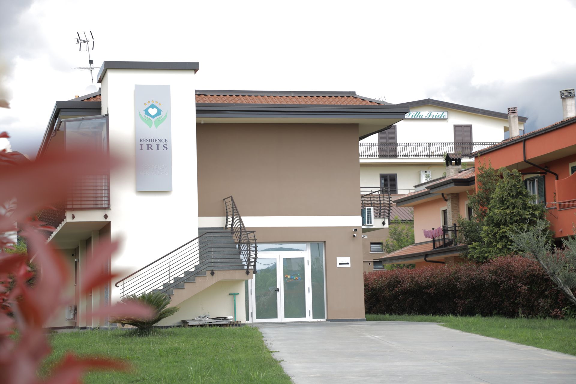 Un edificio a due piani color beige con facciata anteriore bianca, un'insegna centrale, una scala esterna e prati verdi.
