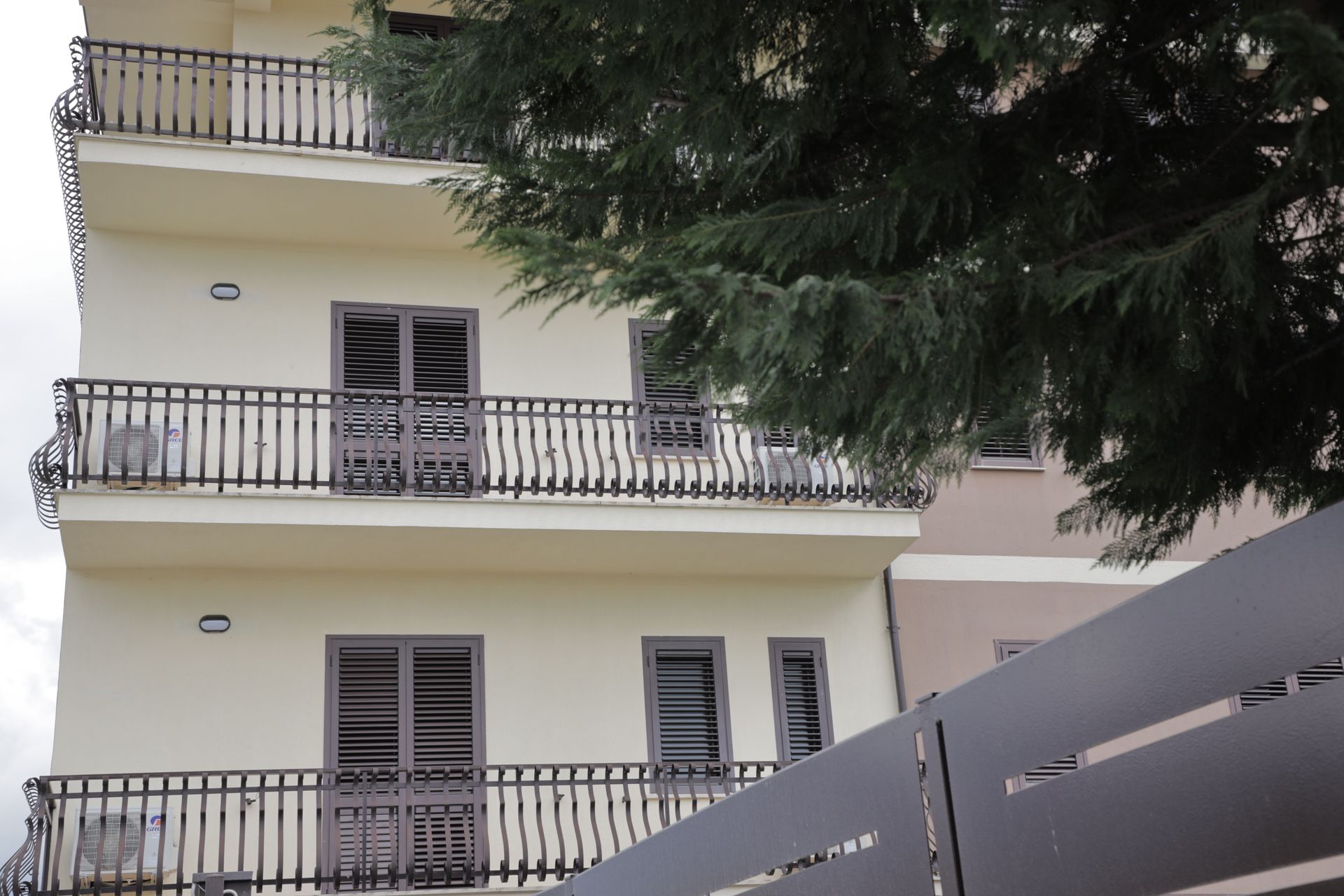 Un condominio a più piani con pareti beige e ringhiere scure, parzialmente nascosto dal ramo di un grande albero sempreverde.