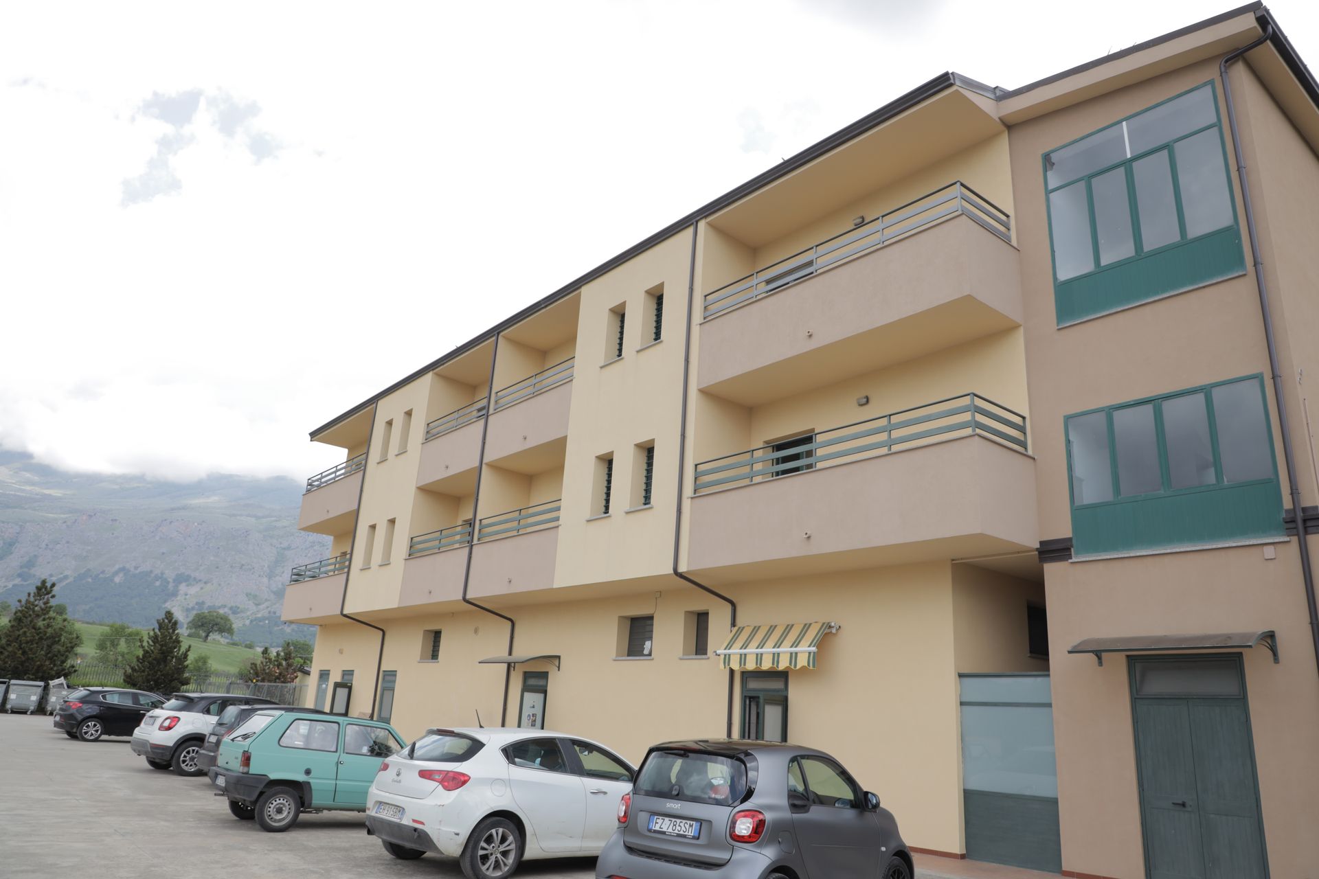 Un condominio beige di tre piani con balconi e un parcheggio con diverse auto, situato di fronte a una montagna.