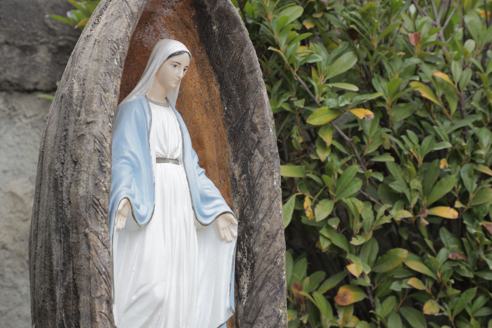 Una statua raffigurante una figura con una veste blu e bianca, in piedi all'interno di un recinto dalla forma a mandorla, accanto a un cespuglio verde.