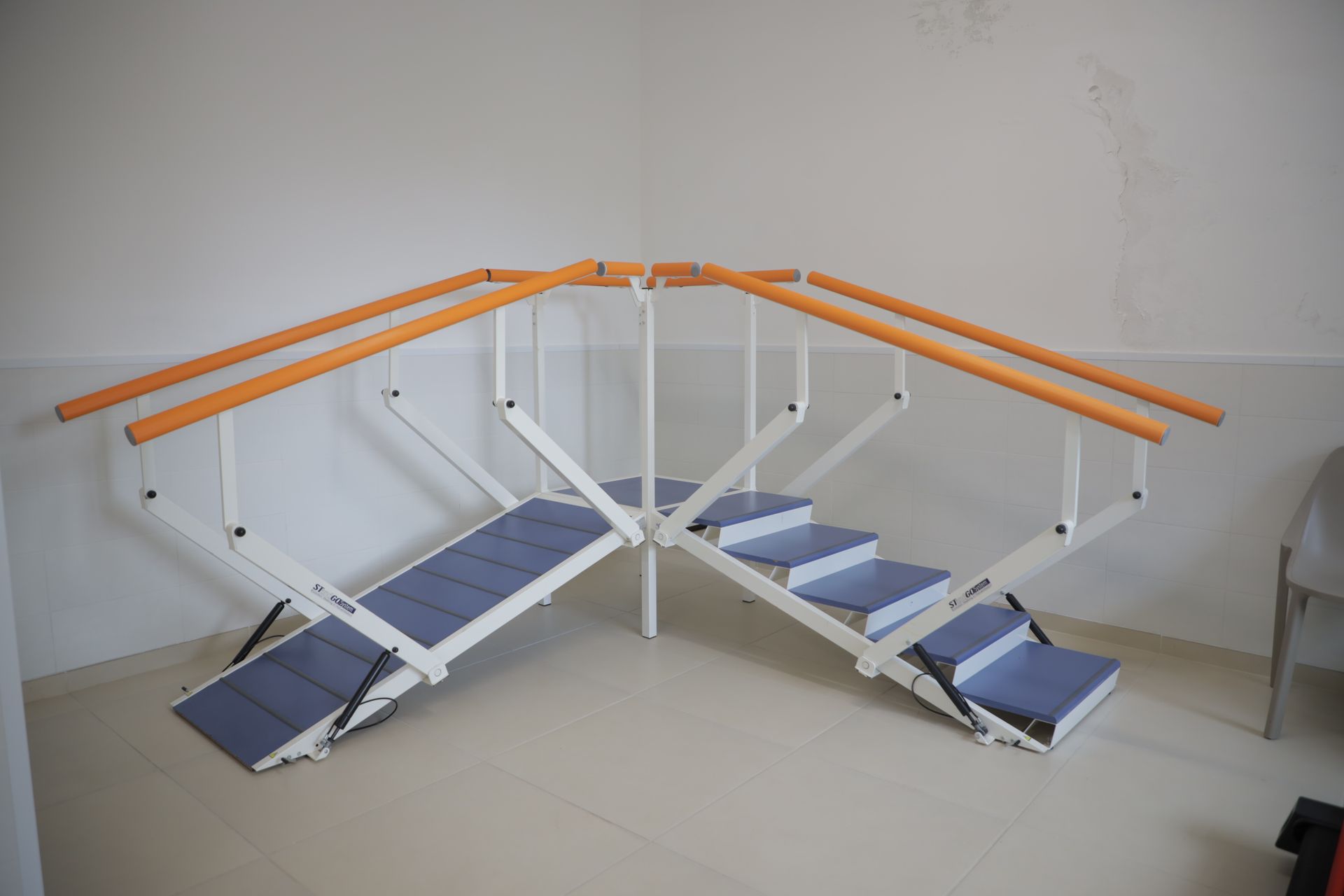 Scala per fisioterapia con corrimano paralleli e rampa integrata, posizionata in un angolo di una stanza clinica.