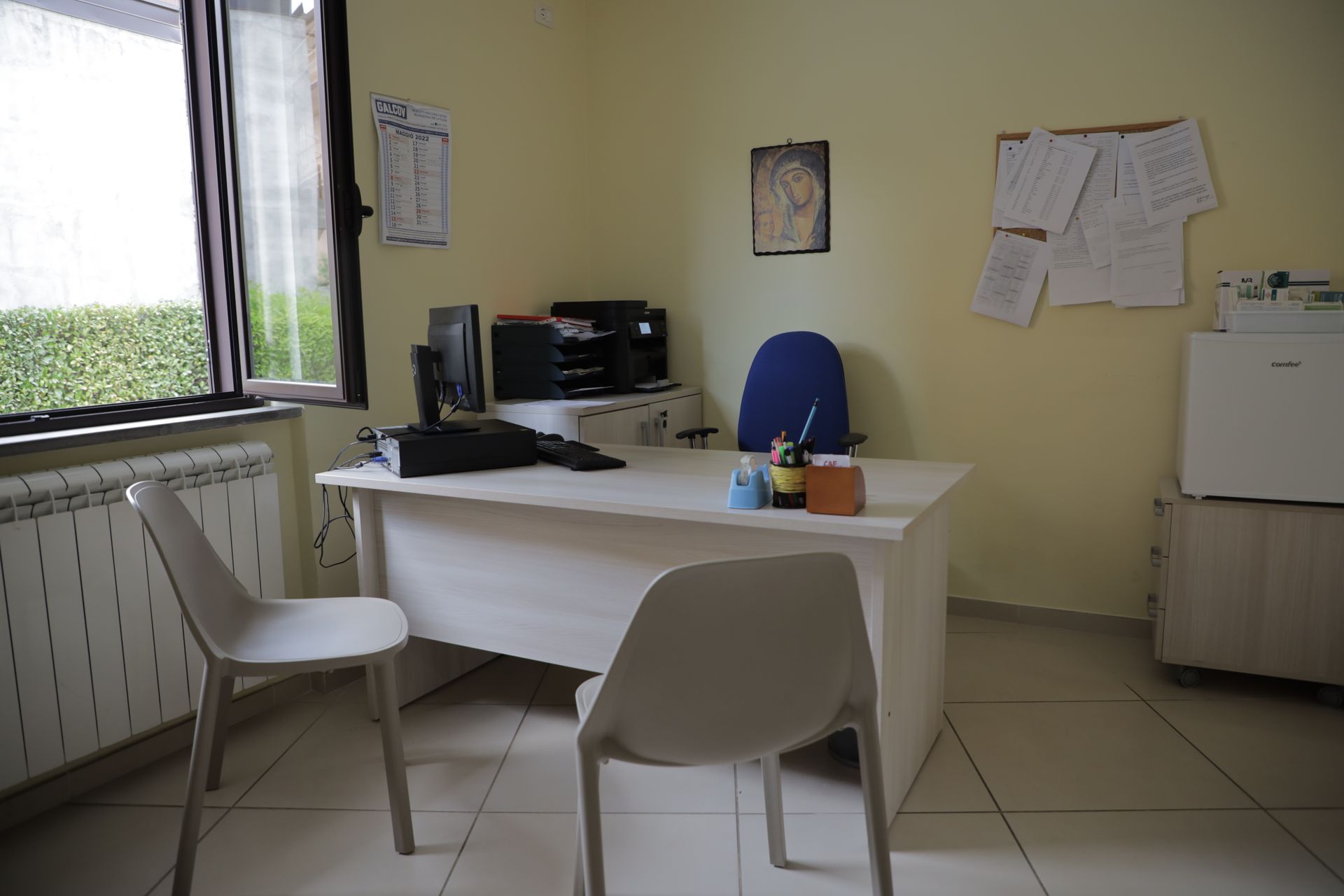 Un piccolo ufficio con una scrivania bianca, un computer, due sedie, un termosifone e fogli appuntati alle pareti giallo pallido.