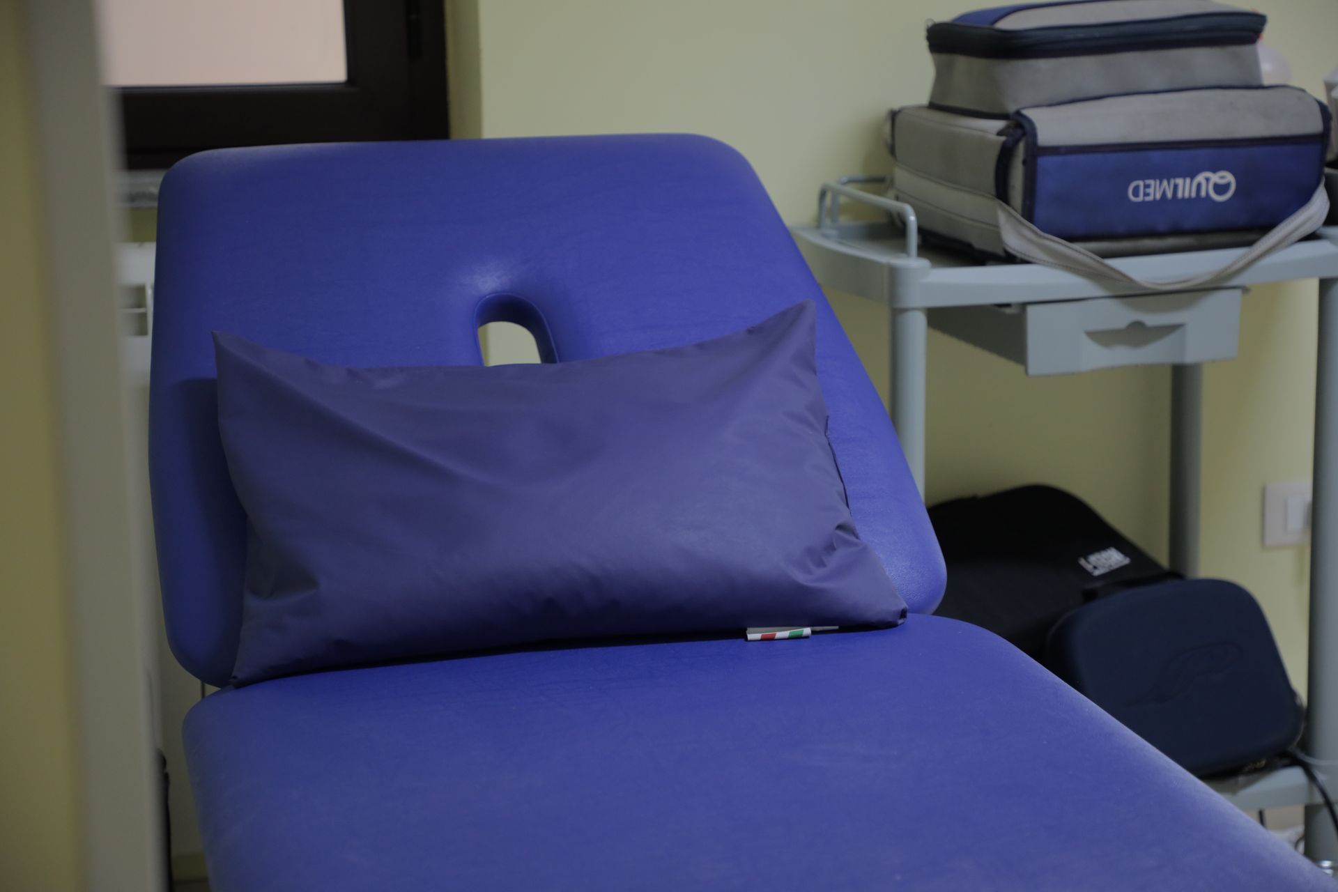 In un ambulatorio, un lettino medico viola con cuscino e un carrello laterale con le attrezzature.