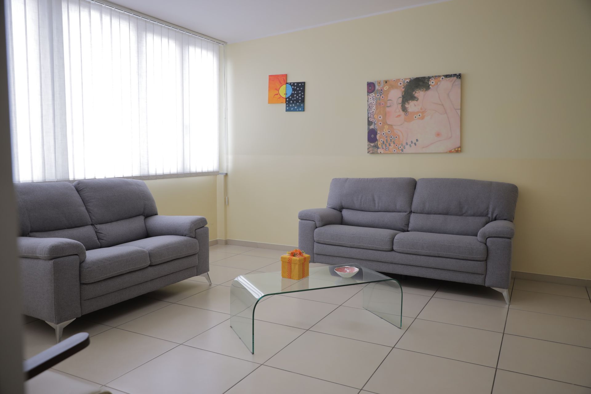 In una stanza dalle pareti chiare con pavimento piastrellato, un tavolino da caffè in vetro e un quadro astratto, due divani grigi sono disposti uno di fronte all'altro.
