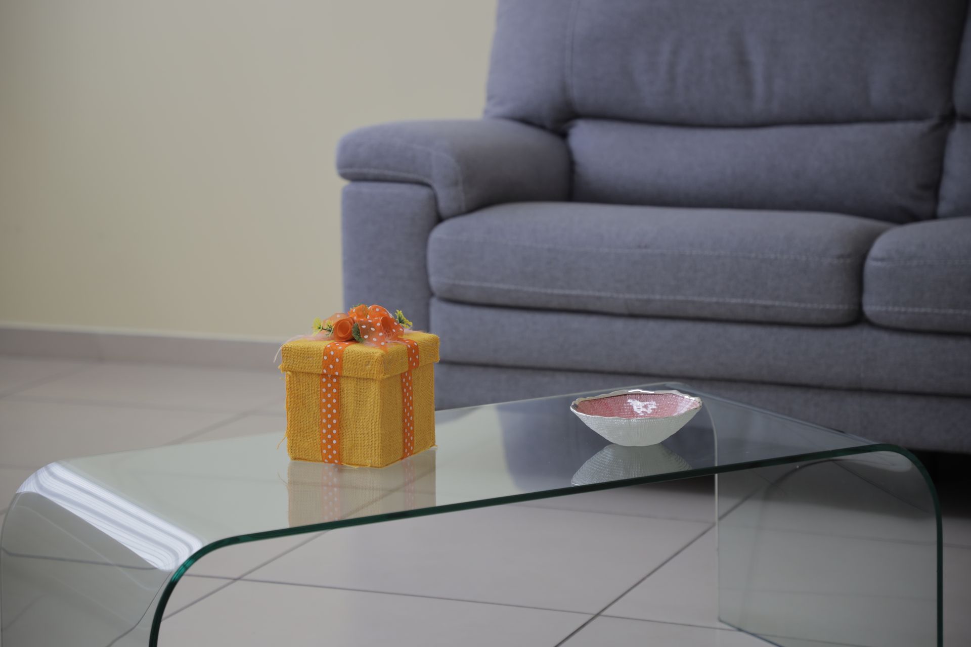 Una scatola regalo gialla con un nastro arancione è appoggiata su un tavolino di vetro davanti a un divano grigio.