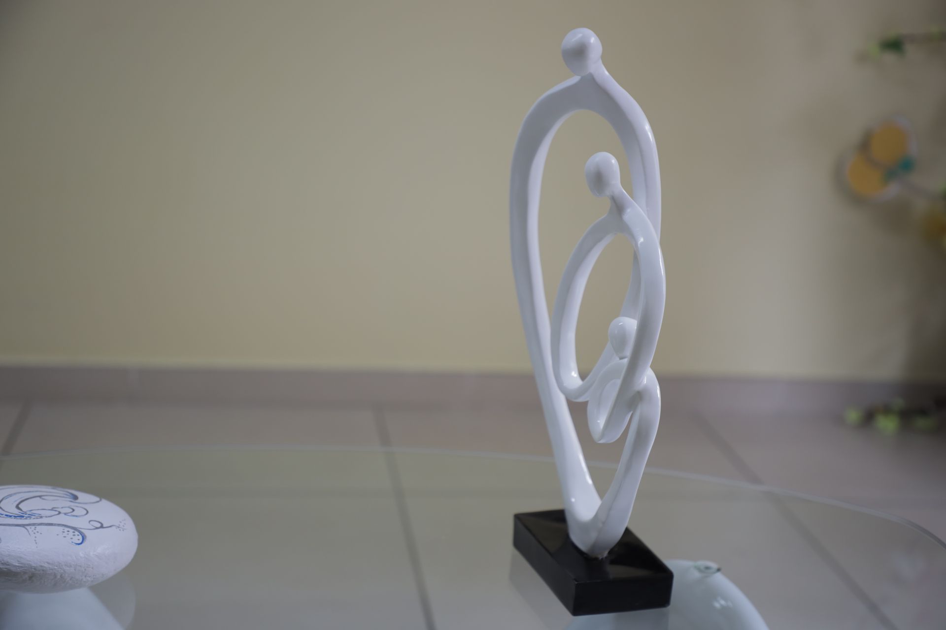Una scultura astratta bianca raffigurante tre figure in un abbraccio, poggiata su una base rettangolare nera su un tavolo di vetro.