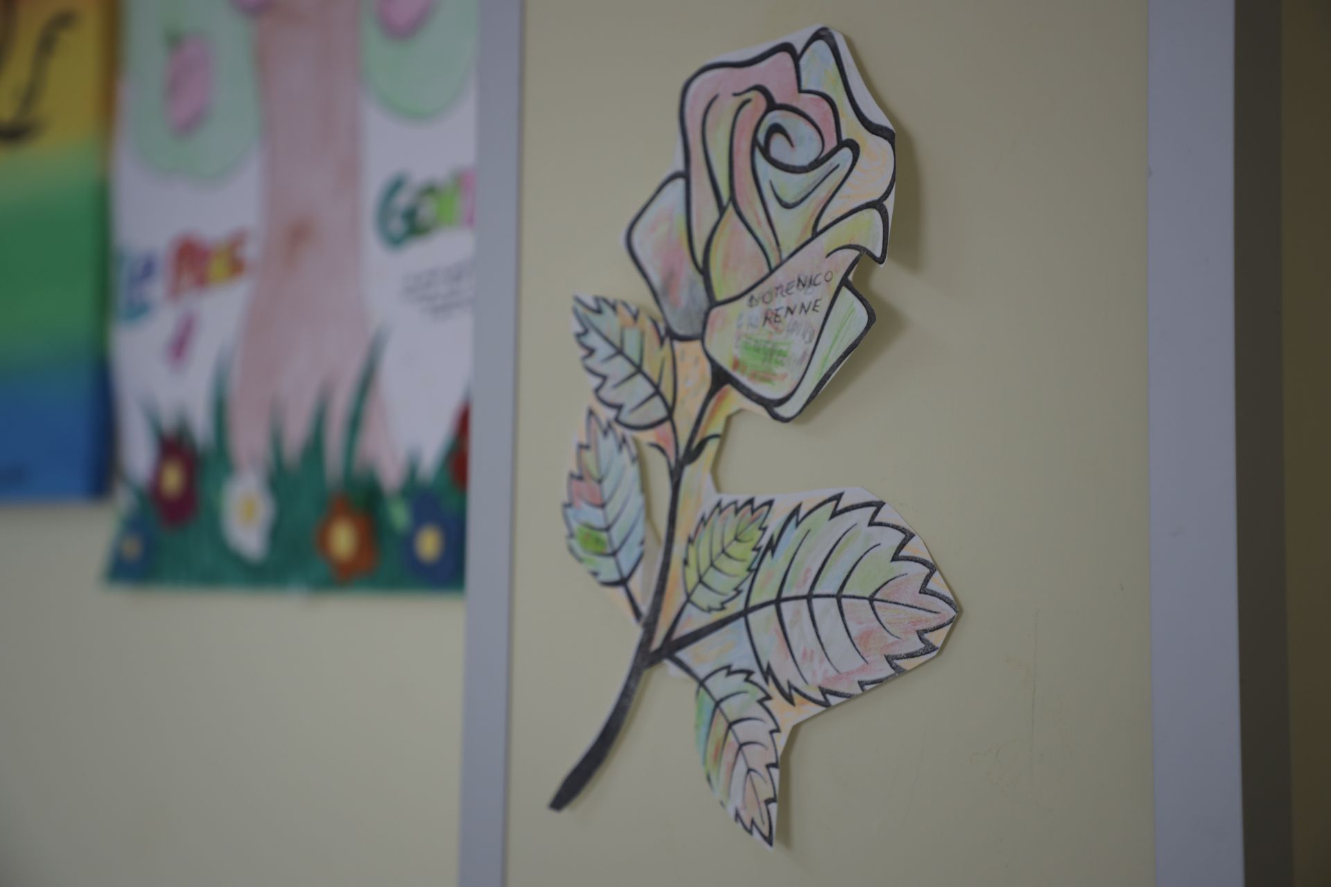 Una rosa di carta ritagliata, dai colori pastello e disegnata a mano, è attaccata a una parete giallo chiaro, con un altro disegno sfocato nelle vicinanze.