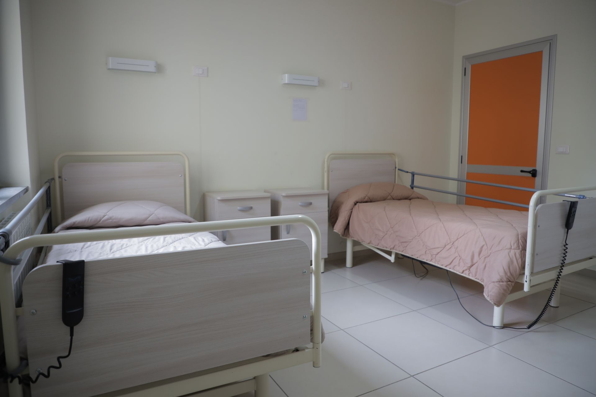Due letti d'ospedale con lenzuola beige e sponde laterali in una stanza con pareti bianche e porta arancione chiaro.