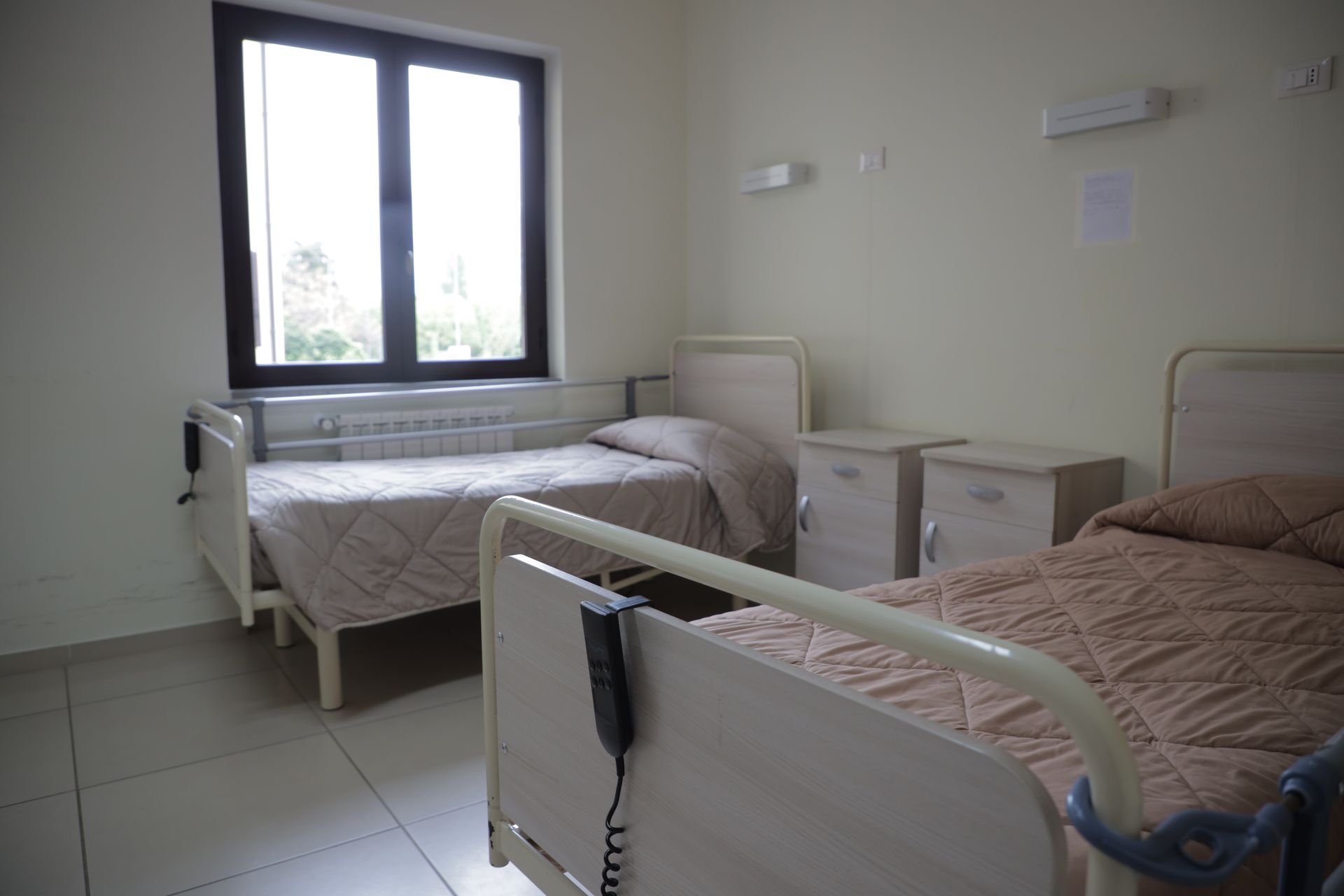 Due letti d'ospedale con lenzuola trapuntate beige in una stanza con finestra e pareti bianche.