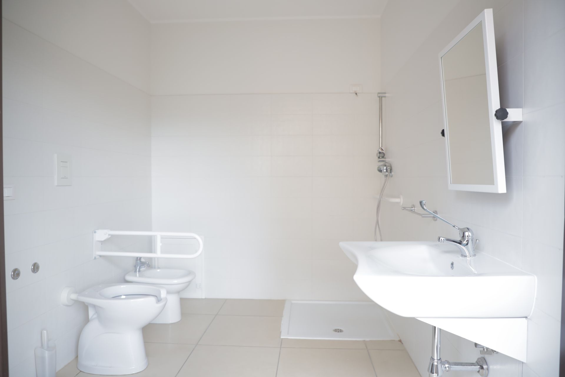 Un bagno accessibile di colore bianco, dotato di WC con maniglione di sostegno, bidet, lavabo a parete e zona doccia.