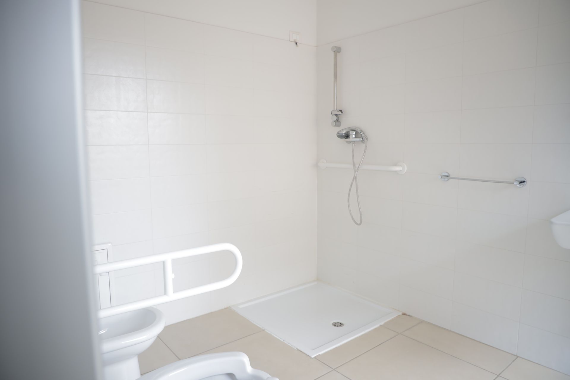 Bagno accessibile con box doccia piastrellato di bianco, maniglioni di sostegno e WC con corrimano.